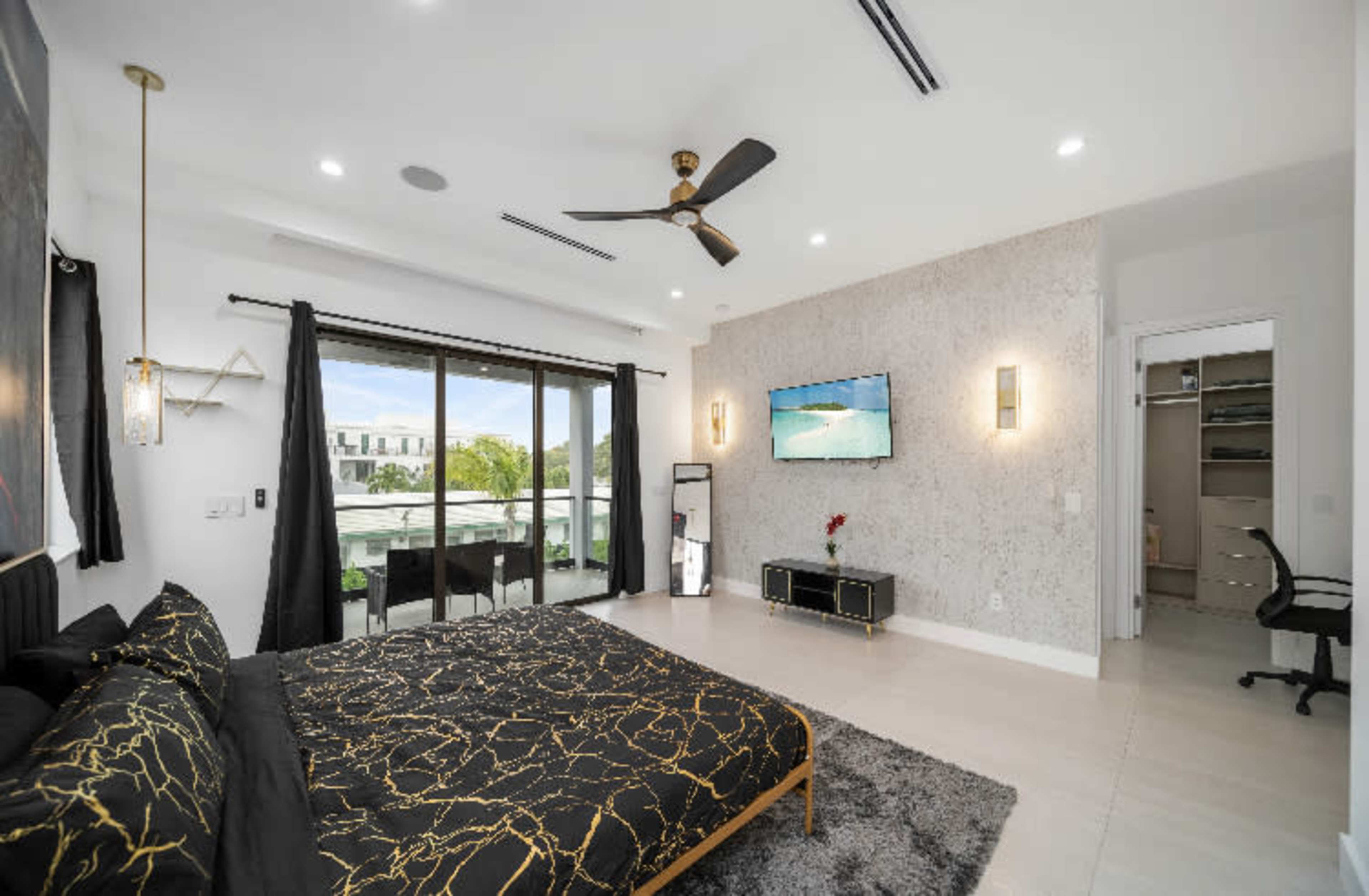 A modern bedroom featuring a large bed with a dark comforter, a sliding glass door leading to a balcony, and a mounted television on the wall.