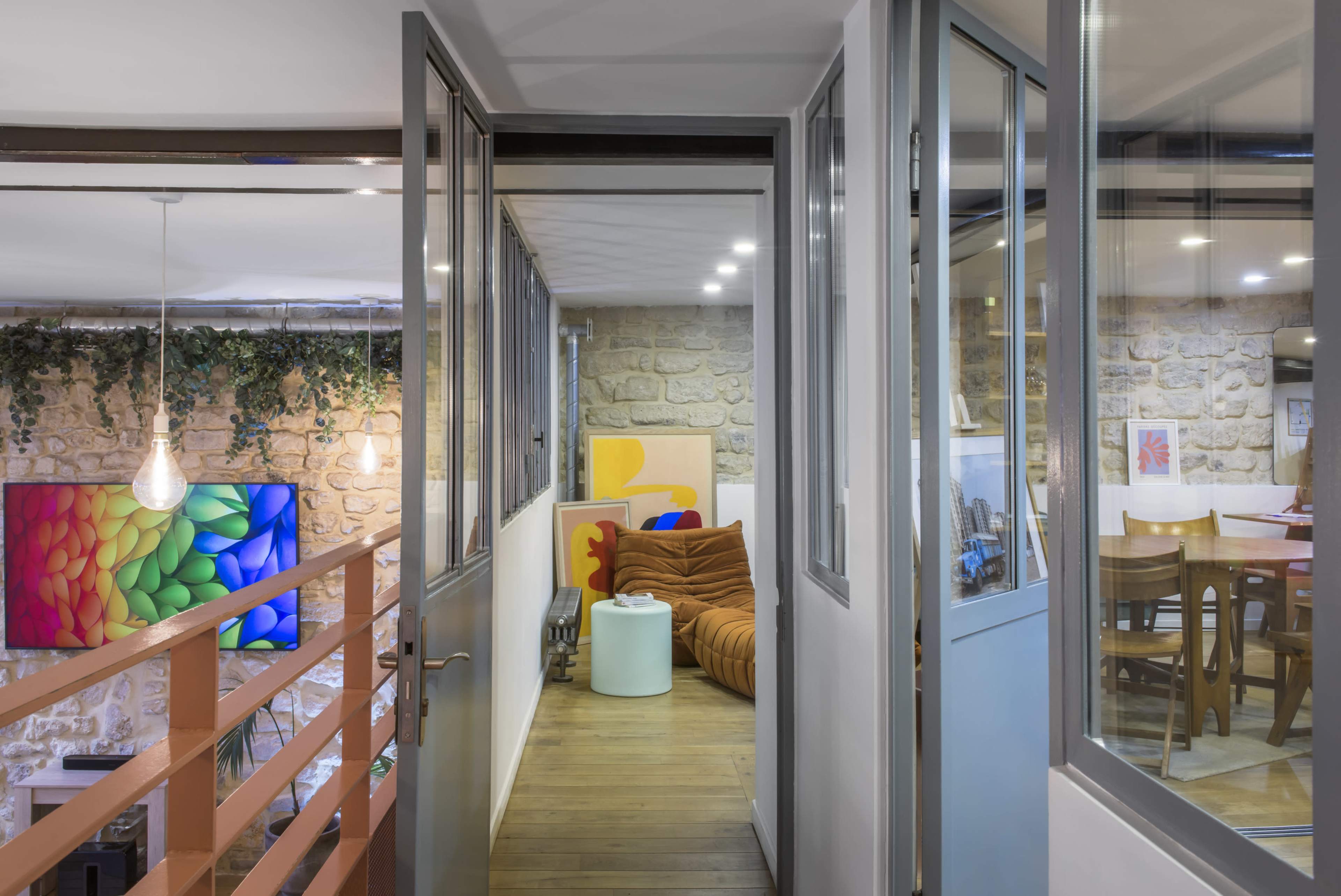 A hallway with glass doors opens to a living space featuring a sofa, a colorful piece of art, and a dining area visible in the background.