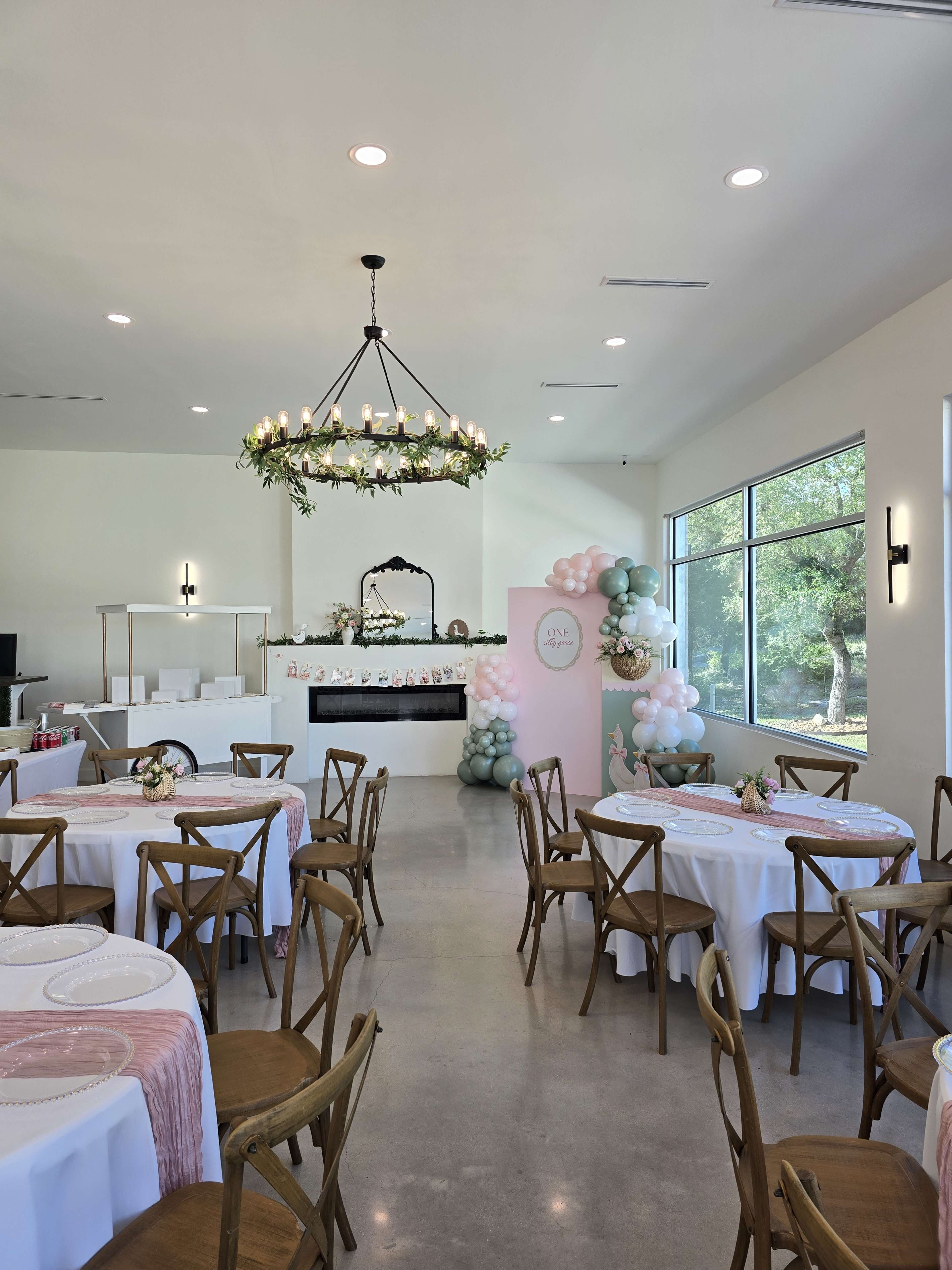 A spacious event room features round tables with white tablecloths, a floral chandelier, and pastel balloon decorations against a wall with greenery.