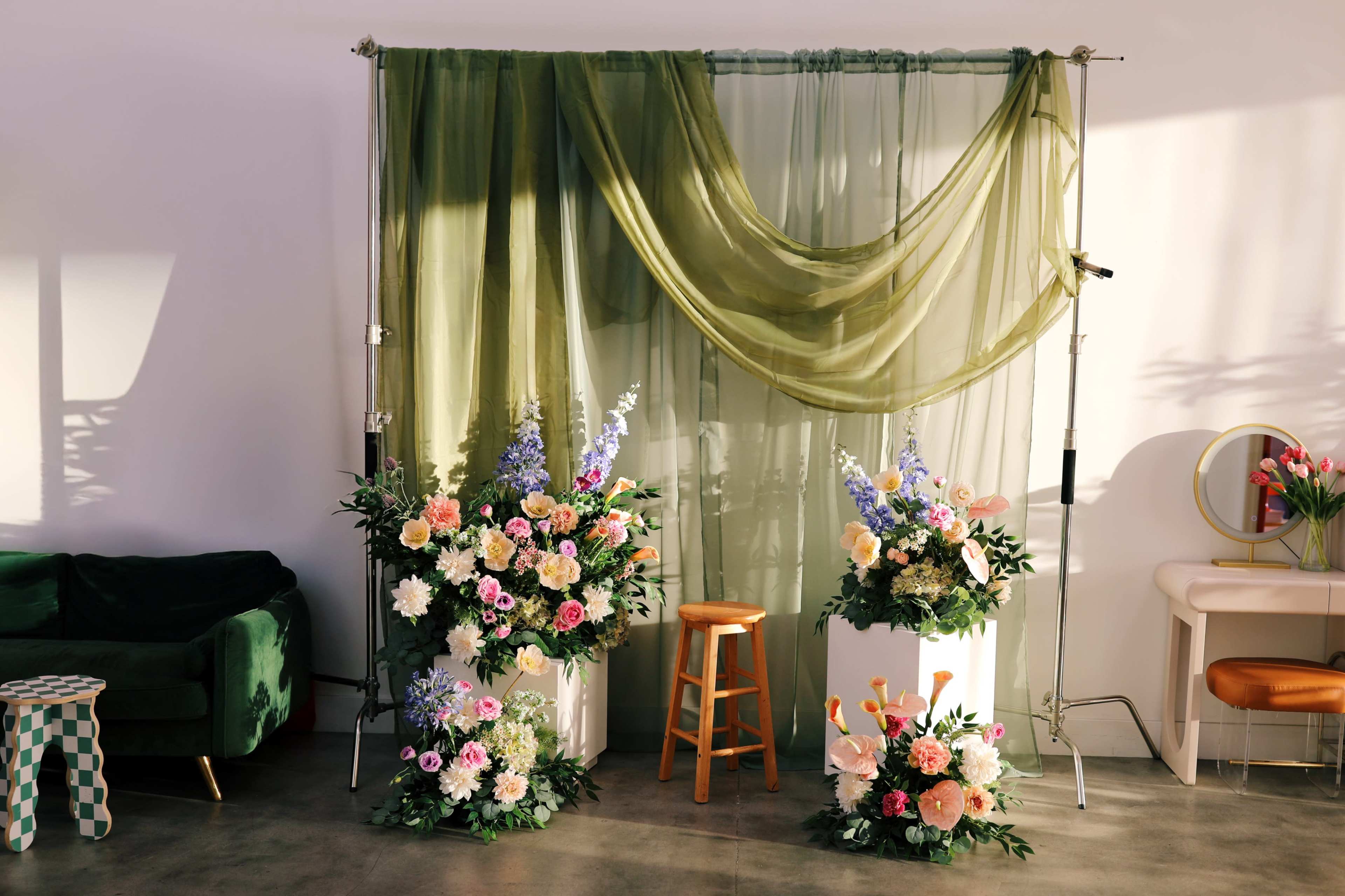 A floral backdrop with draped green fabric and two flower arrangements is set in a modern interior space featuring a green couch and a wooden stool.