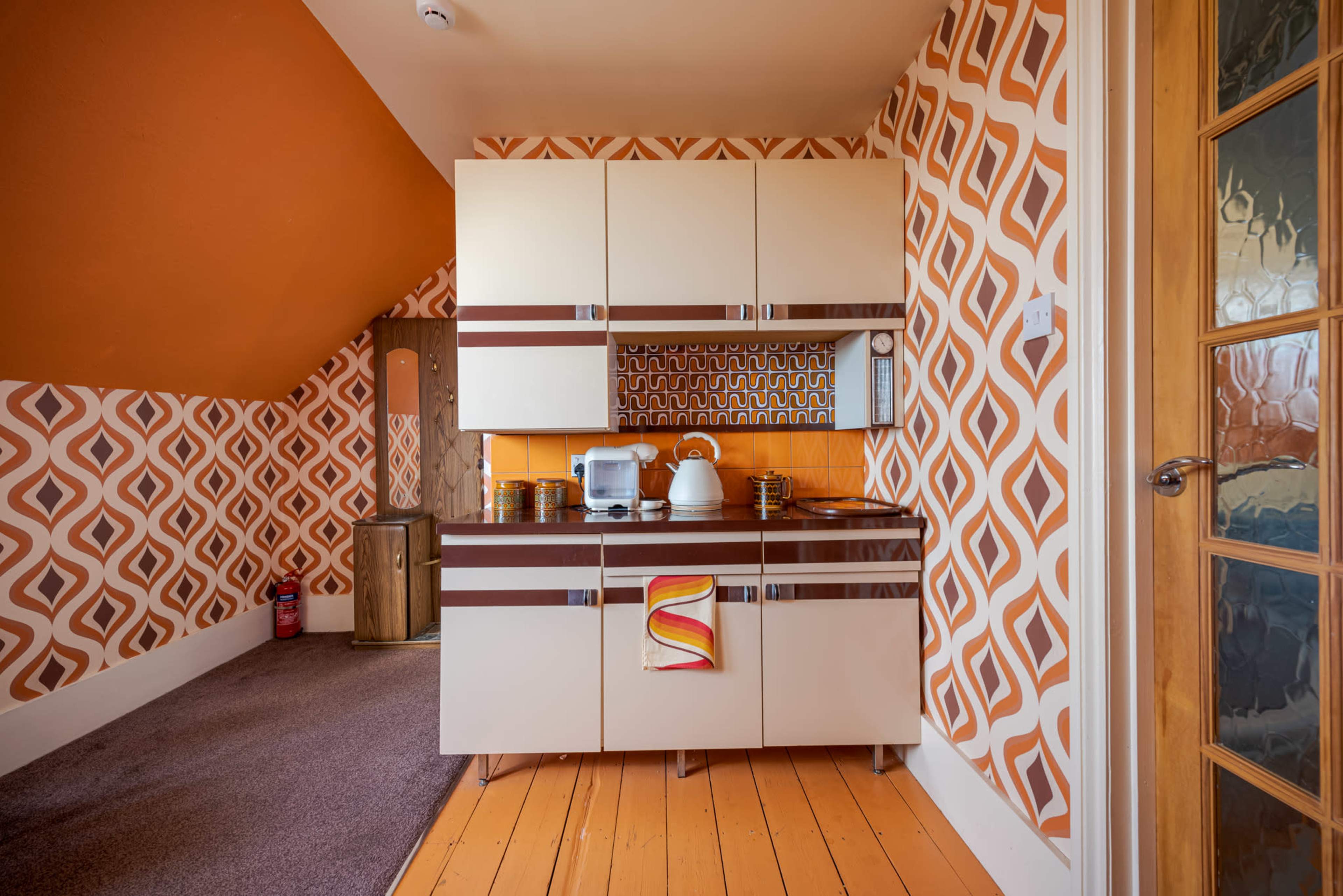 The kitchen features retro-style cabinetry with a patterned wallpaper in orange and brown tones, accompanied by wooden flooring.