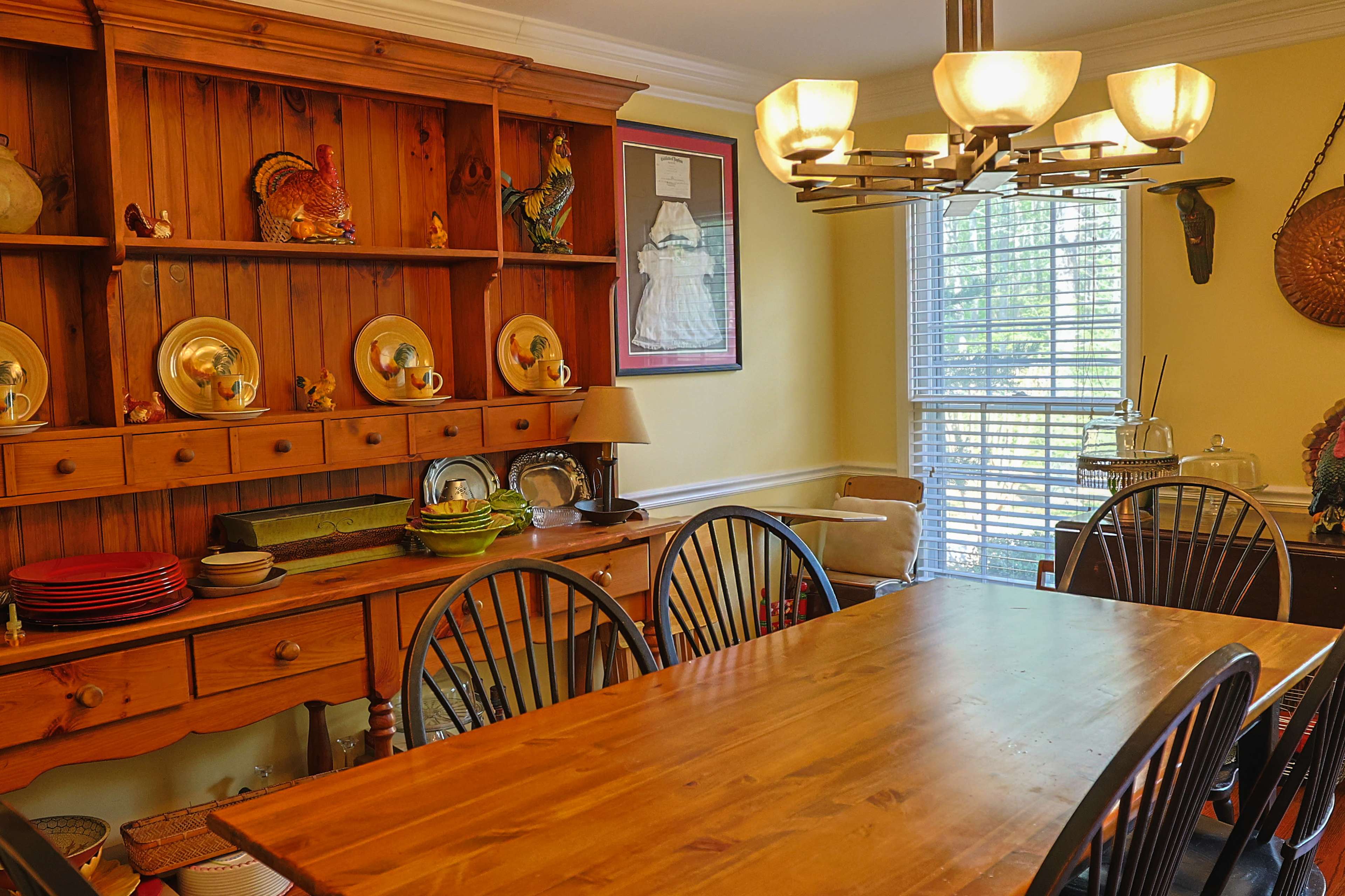 The image features a dining room with a wooden table surrounded by black chairs, a hutch displaying dishes and decor, and a large window letting in natural light.
