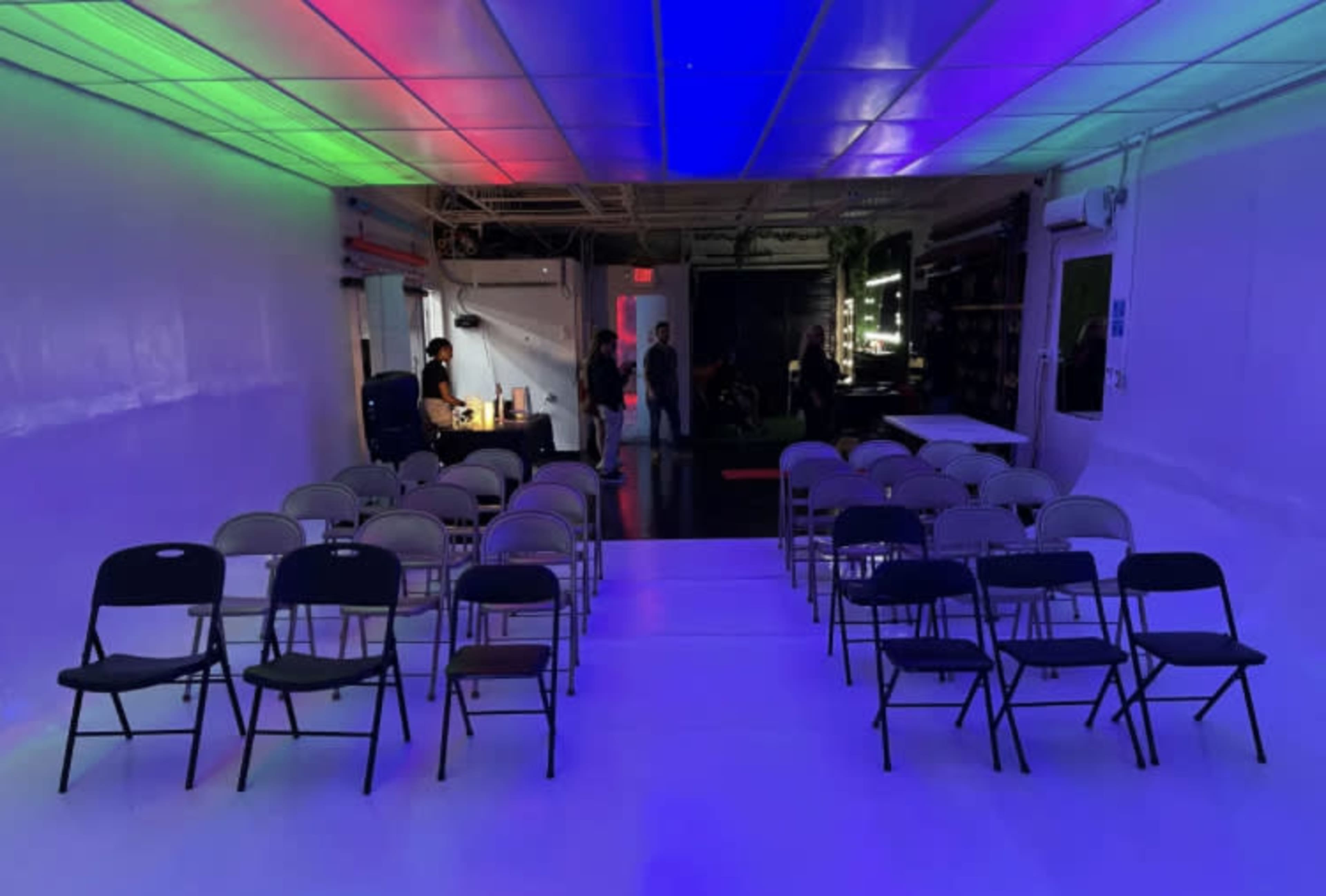 The image shows a brightly lit, empty room arranged with folding chairs facing a stage, with a colorful light display in the ceiling.