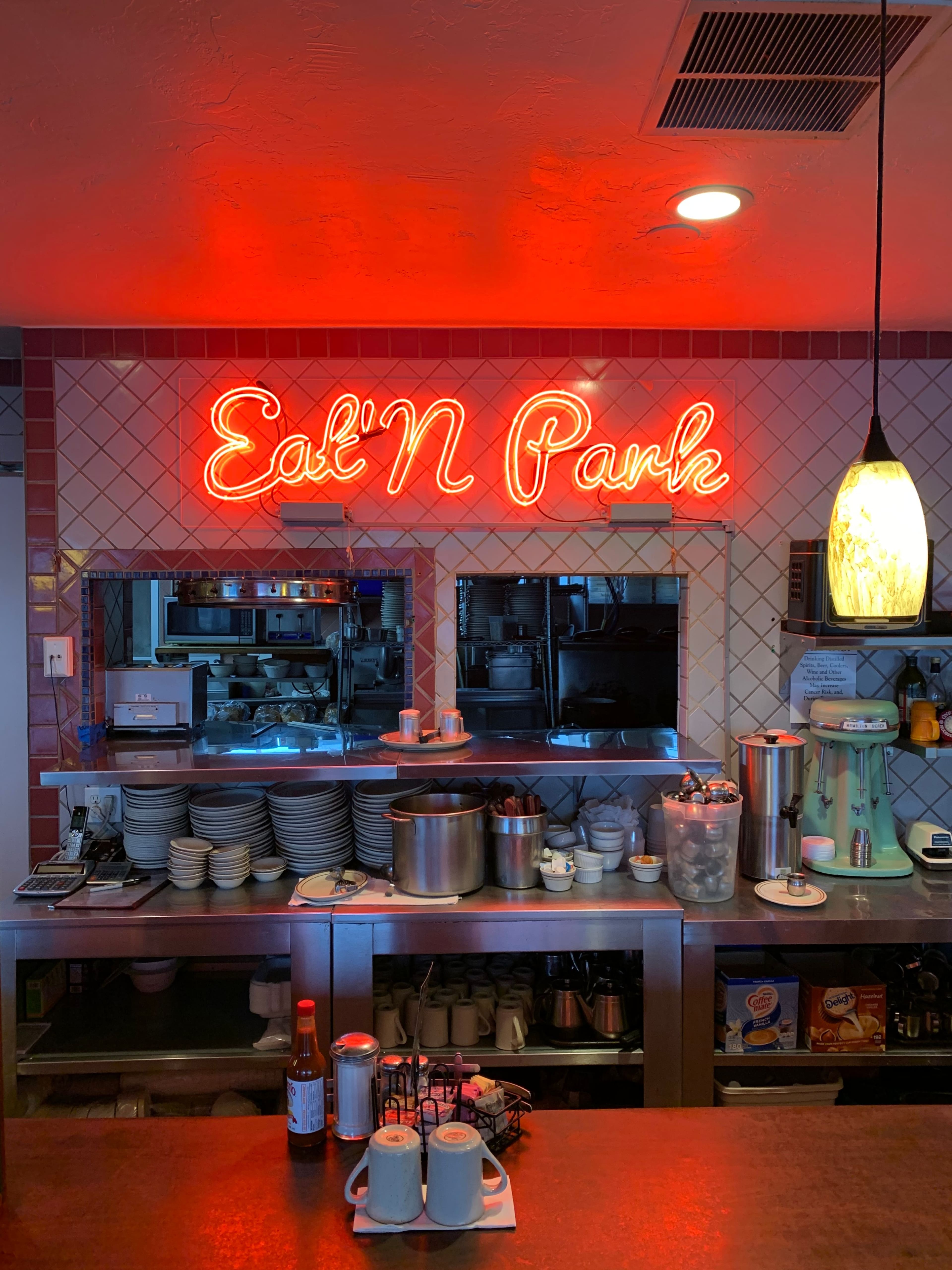 A neon sign reading "Eat'N Park" is illuminated above a restaurant counter with clean plates and various kitchen appliances visible in the background.