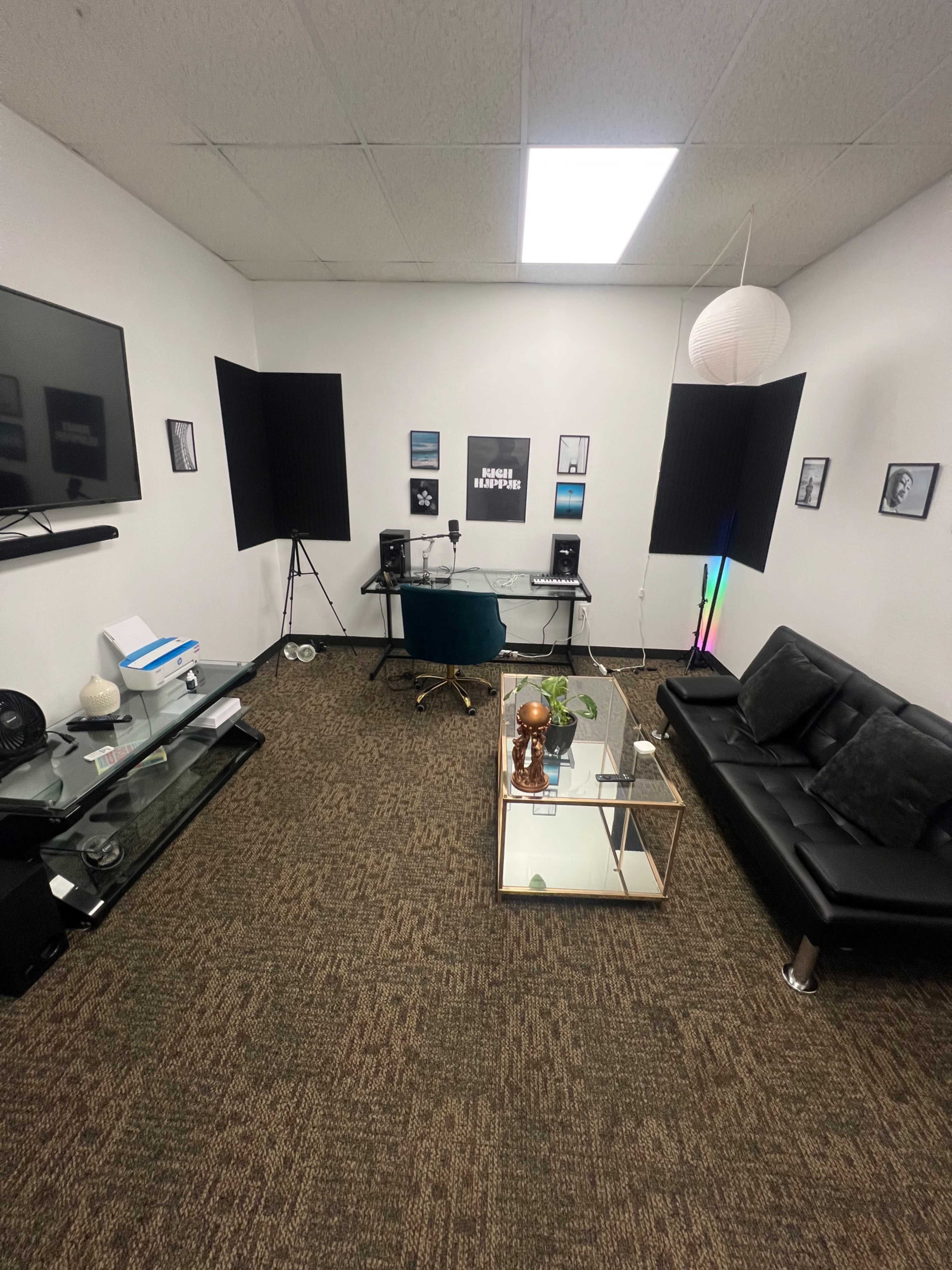 The image shows a modern recording studio with a glass table, a black couch, and soundproofing panels on the walls.