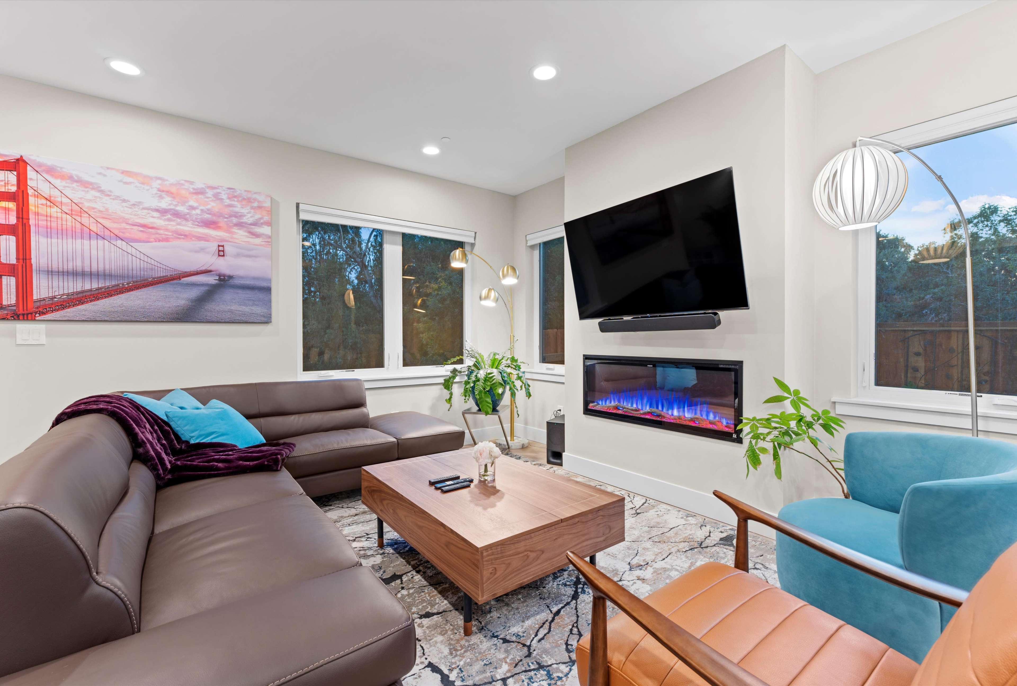 The living room features a sectional sofa, a modern coffee table, a mounted TV, and a wall-mounted electric fireplace, with large windows providing natural light.