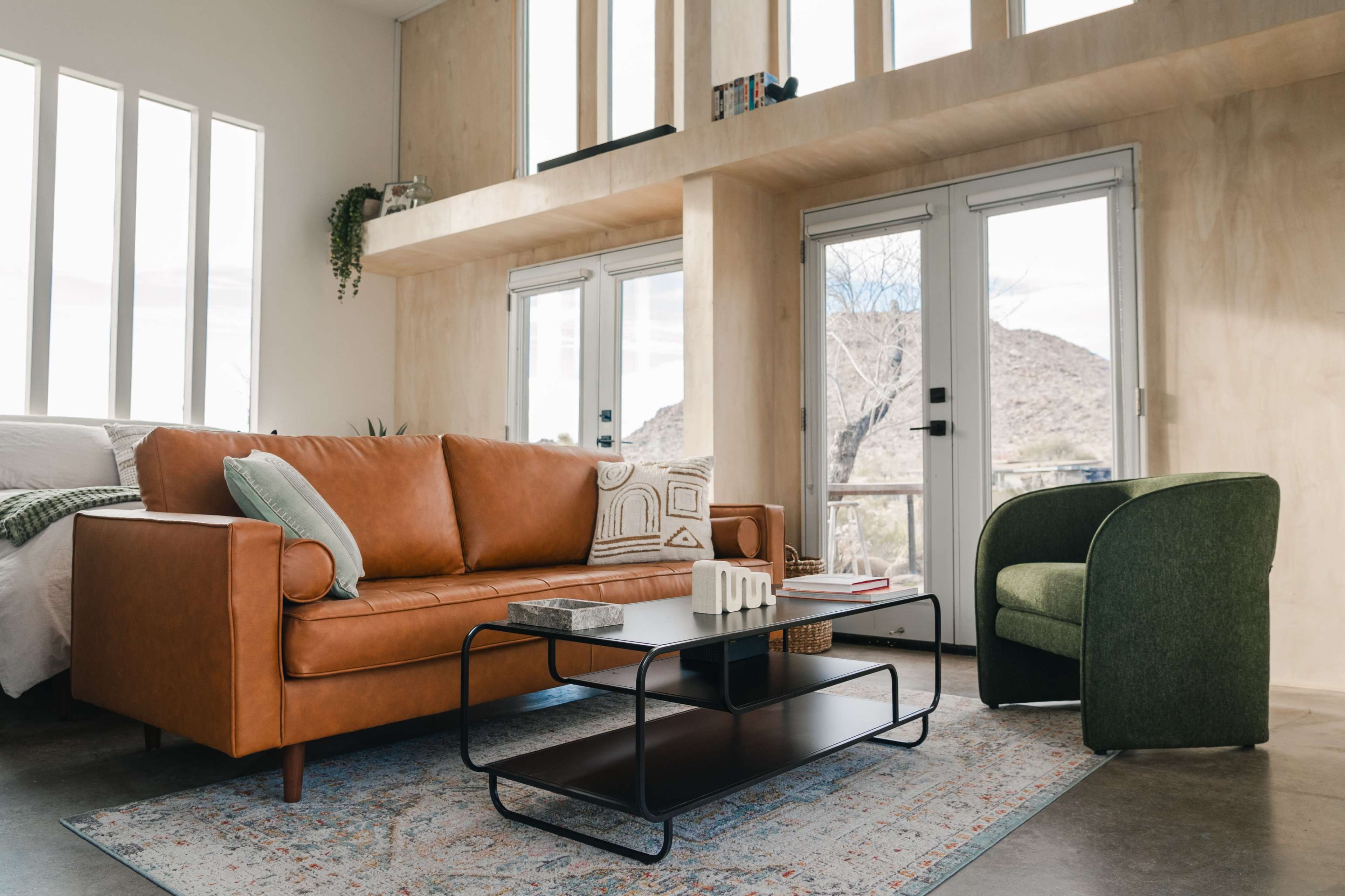 The image shows a modern living area featuring a leather sofa, a green chair, a coffee table with books, and large windows letting in natural light.