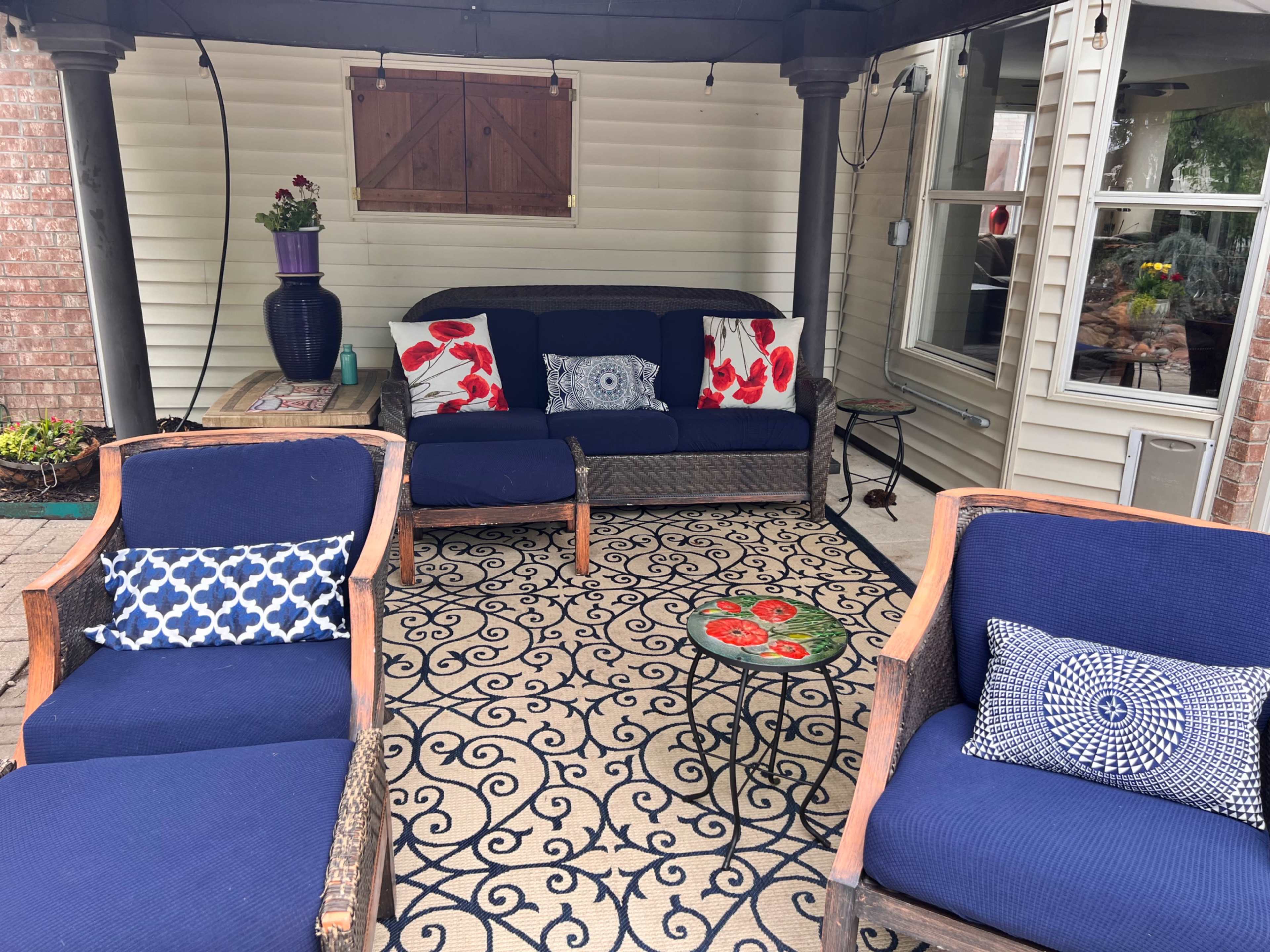 A covered patio featuring a sofa and two armchairs with blue cushions, a small round table, and decorative throw pillows, surrounded by brick walls and a window.