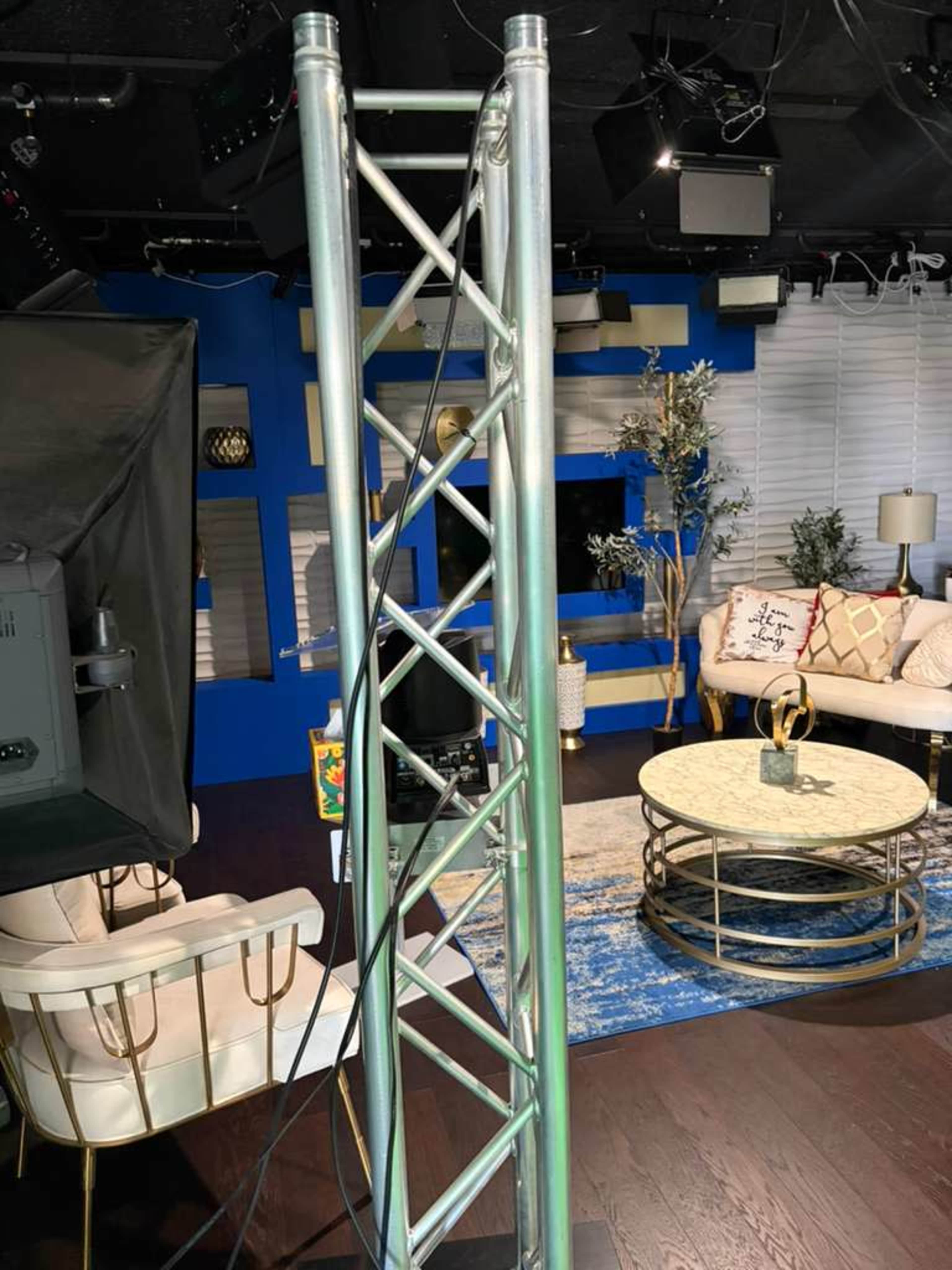 The image shows a studio setup featuring a metal truss structure, cables, and a furnished seating area with a coffee table and decorative elements.