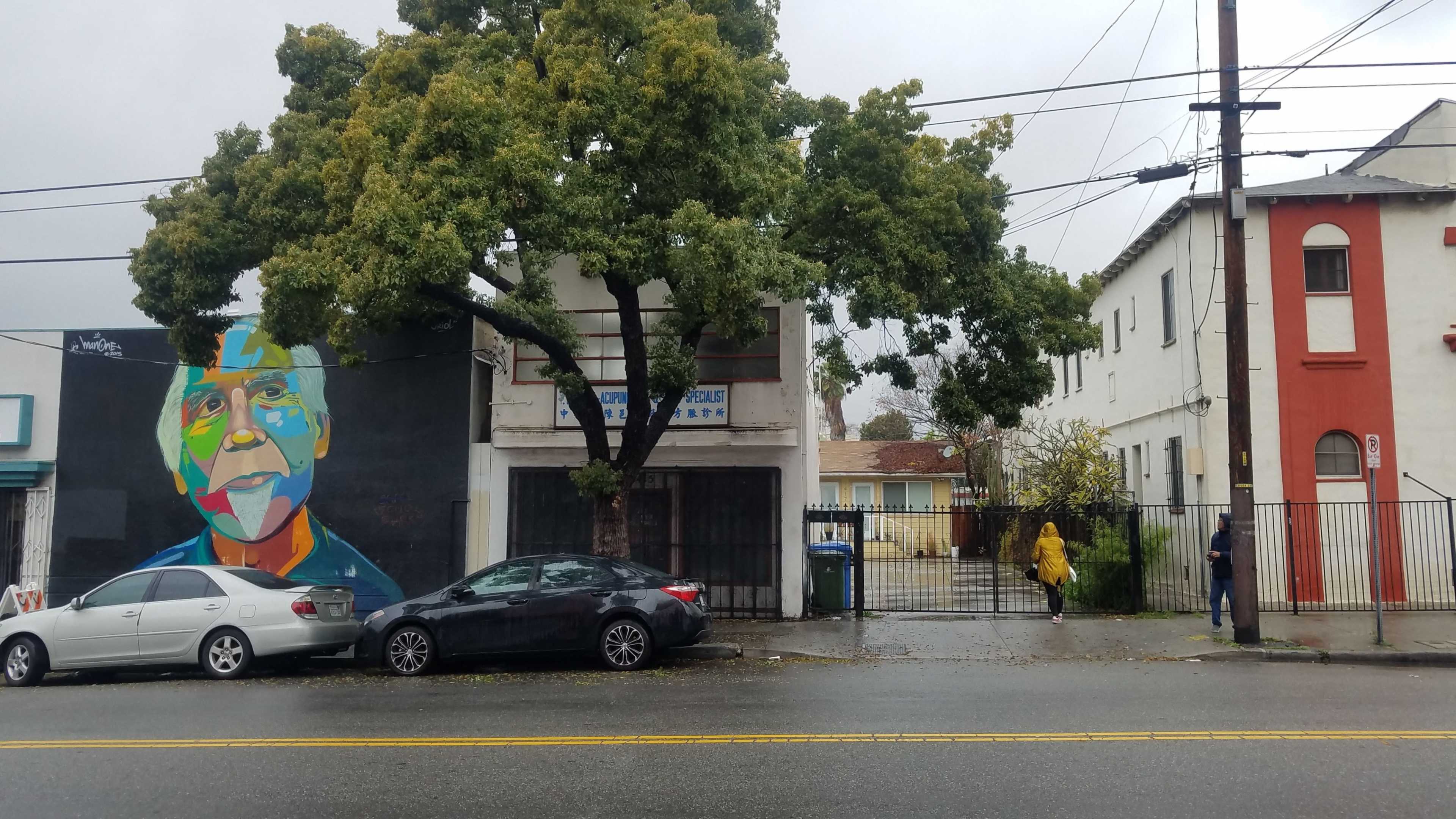 A colorful mural of a man's face is painted on a building next to a street lined with parked cars, while a person in a yellow raincoat walks along the sidewalk in rainy weather.