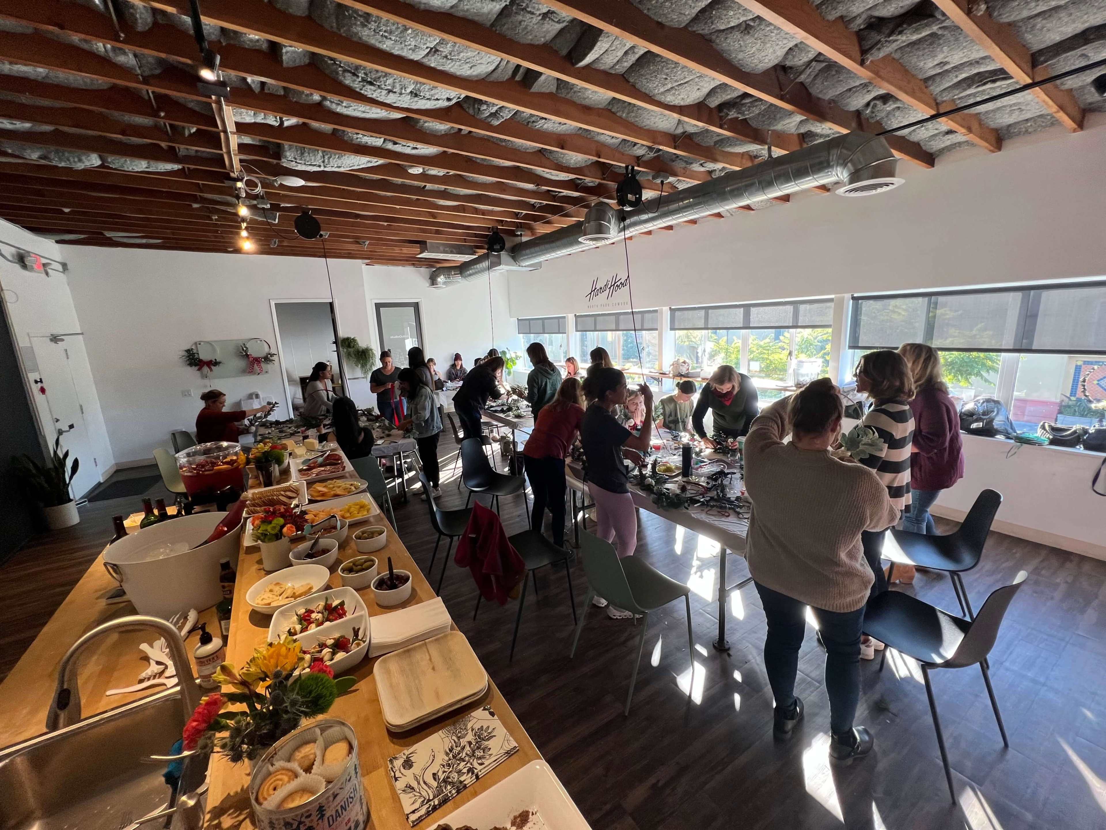 A group of people is gathered around tables in a bright, modern space, preparing food and engaging in activities.