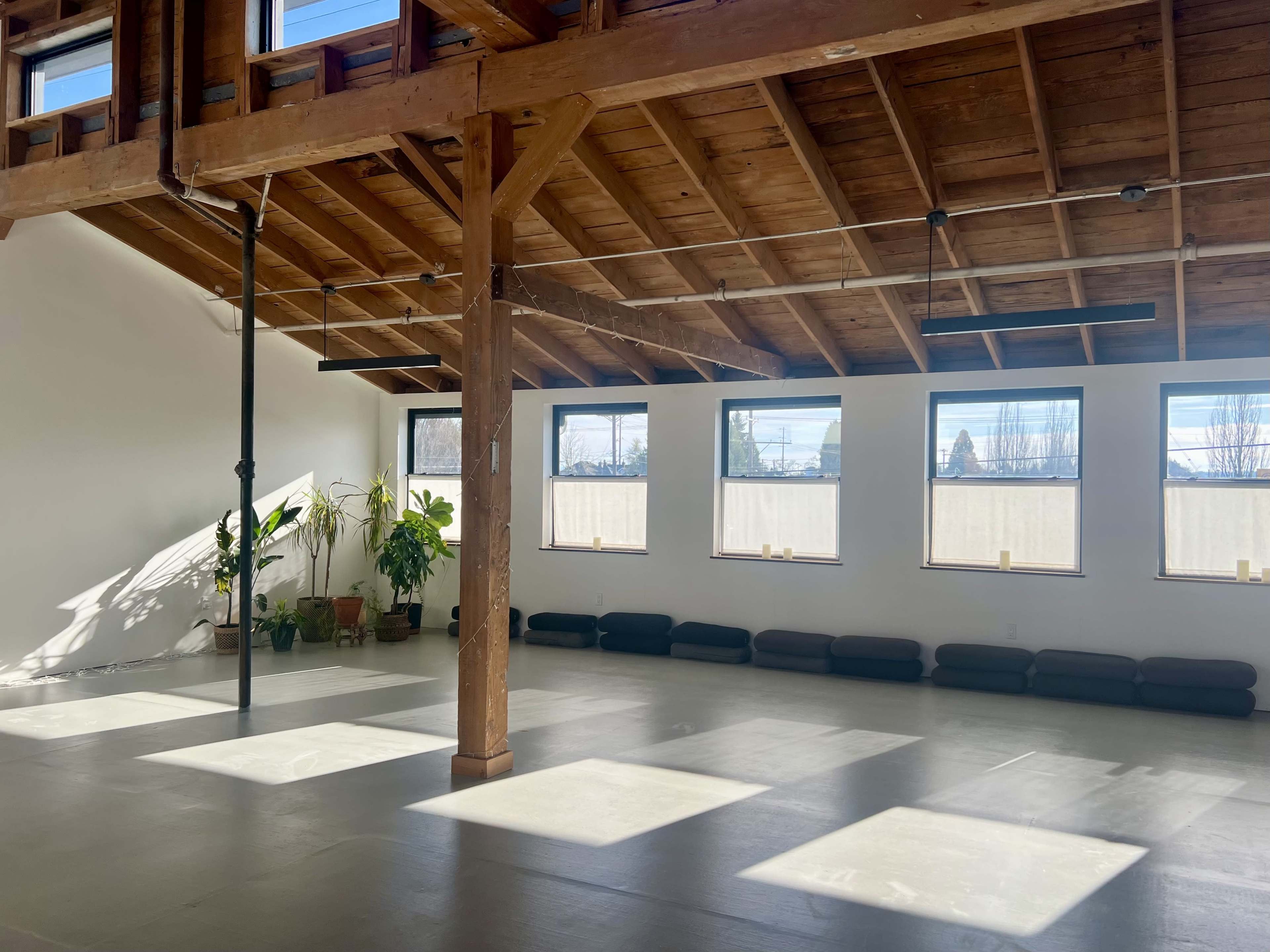 The image shows a spacious indoor area with wooden beams, large windows, and floor cushions arranged along the wall.