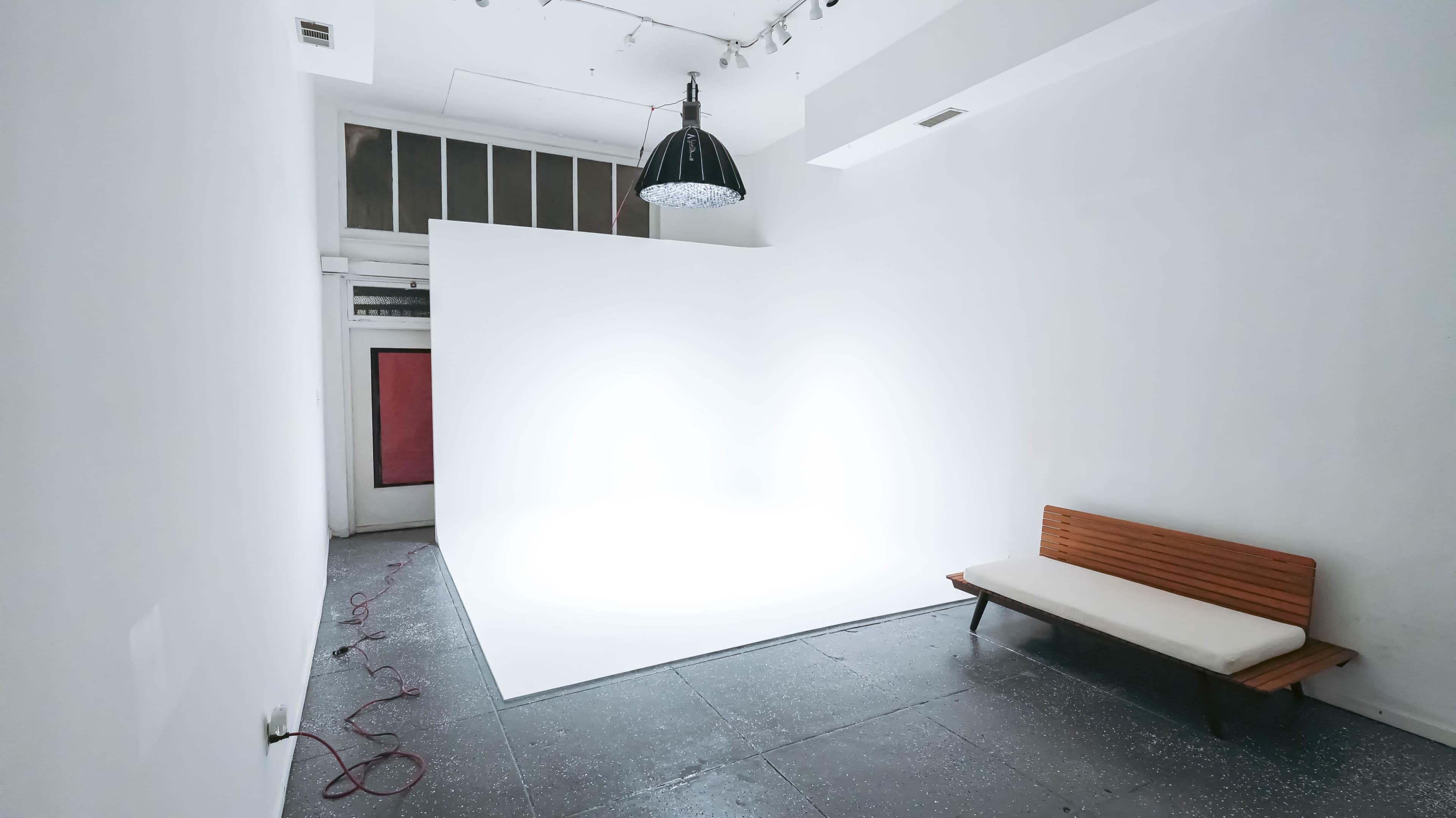 A minimalist interior features a bright white backdrop, a wooden bench, and a pendant light in a spacious room.