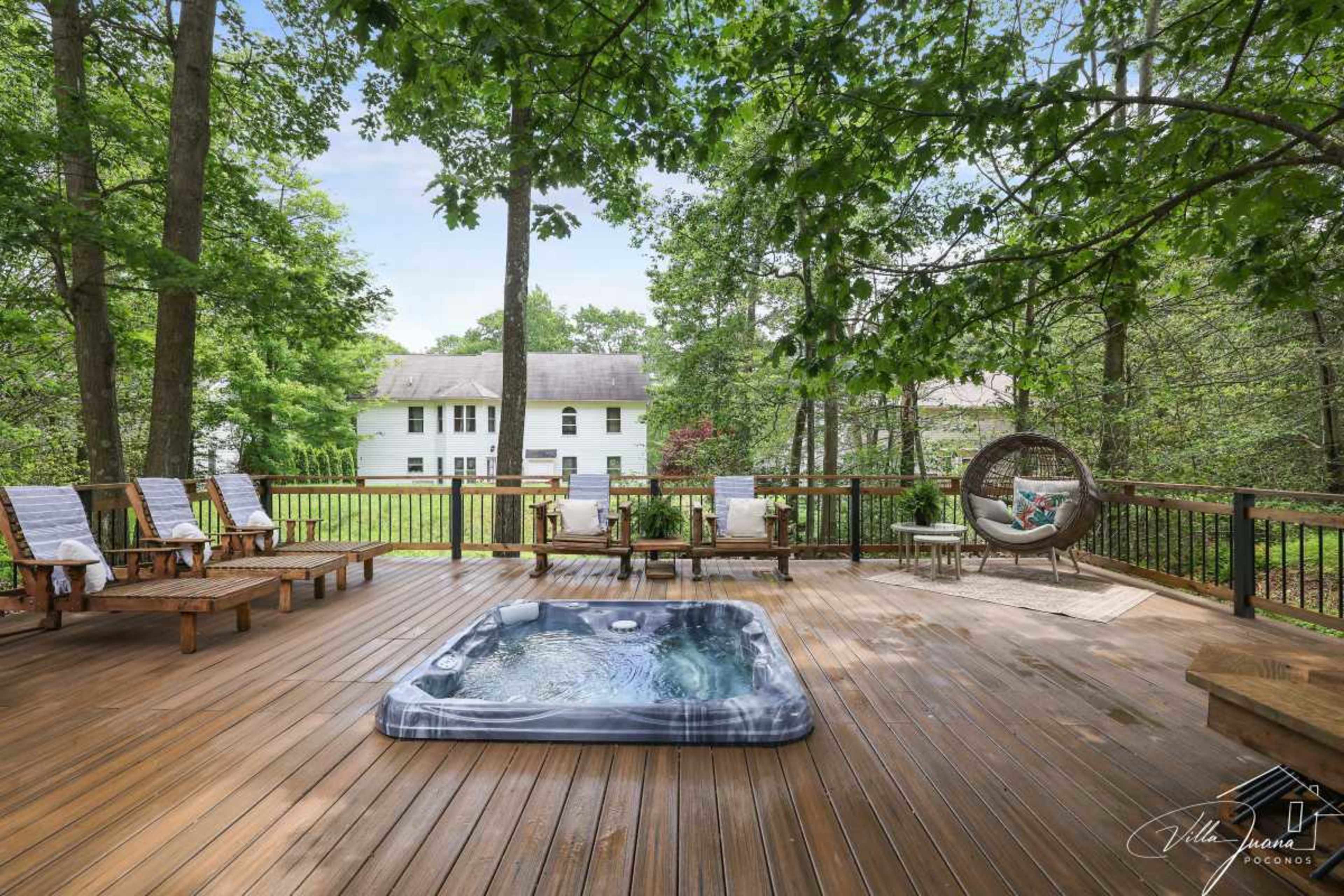 The image shows a wooden deck featuring a hot tub, lounge chairs, and a hanging chair, surrounded by trees and a view of a house in the background.