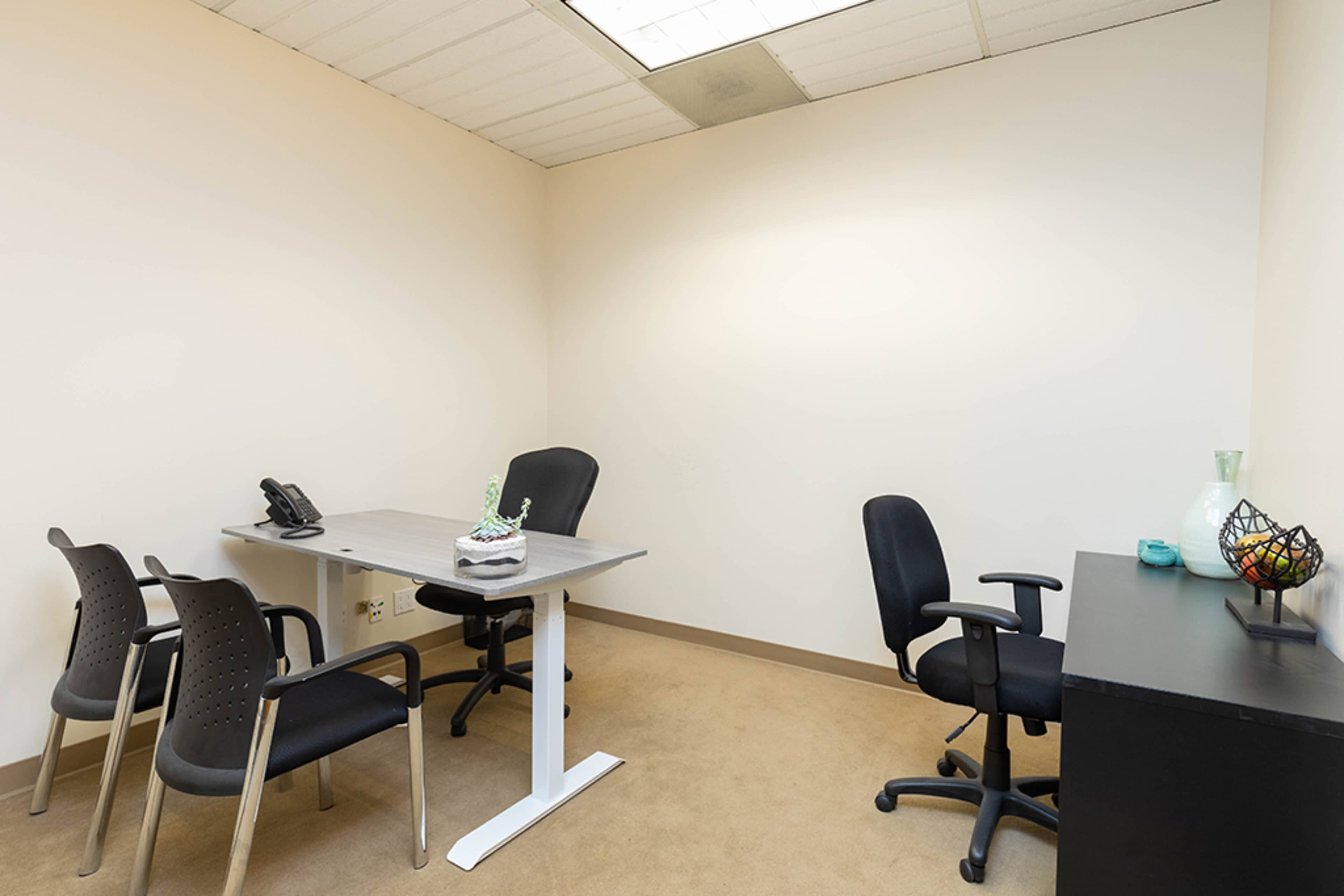 An empty office features a desk with a phone and a small decorative item, four chairs, and a black cabinet along the wall.