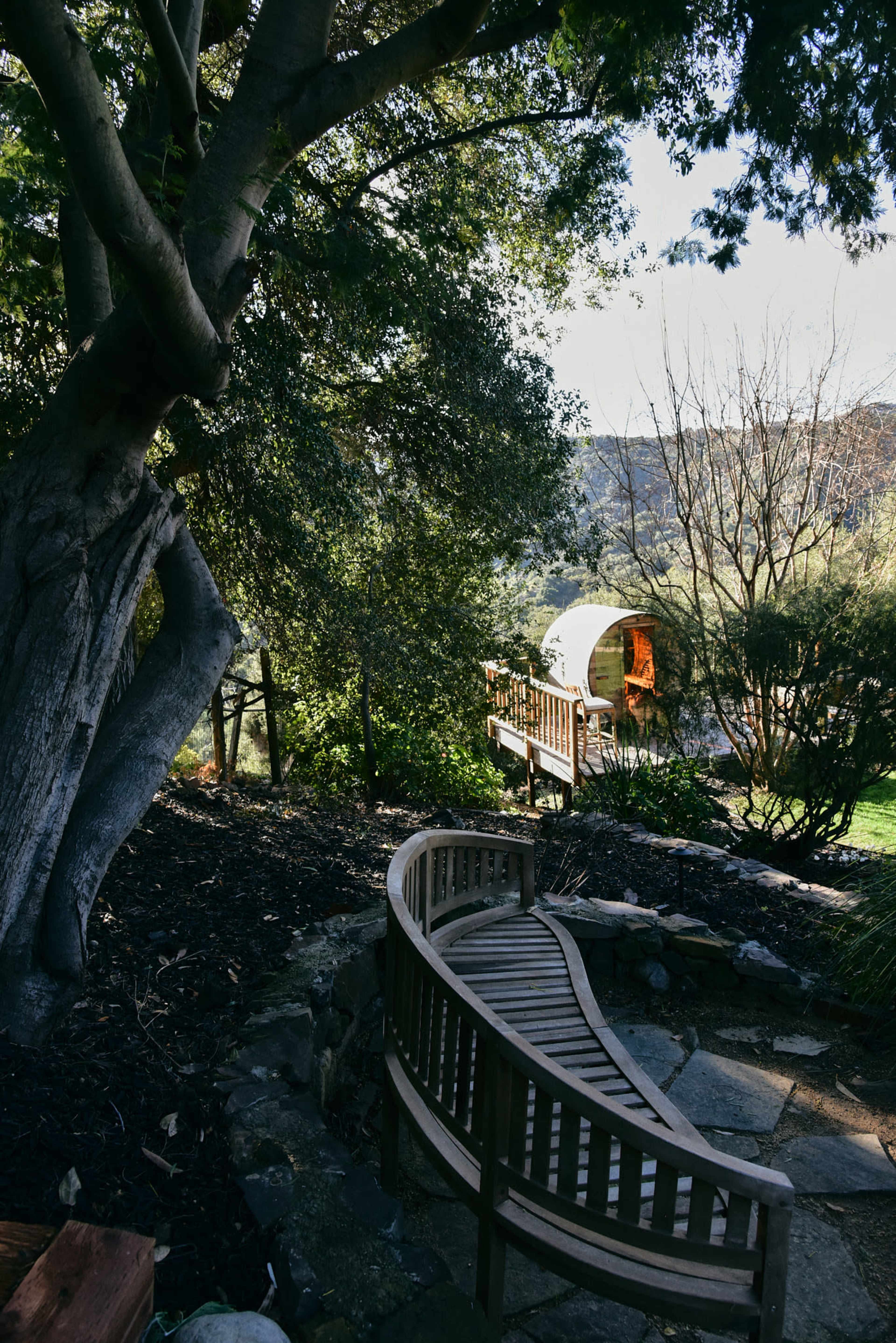 A curved wooden bench is positioned along a stone path, with a view of a wagon and greenery in the background.