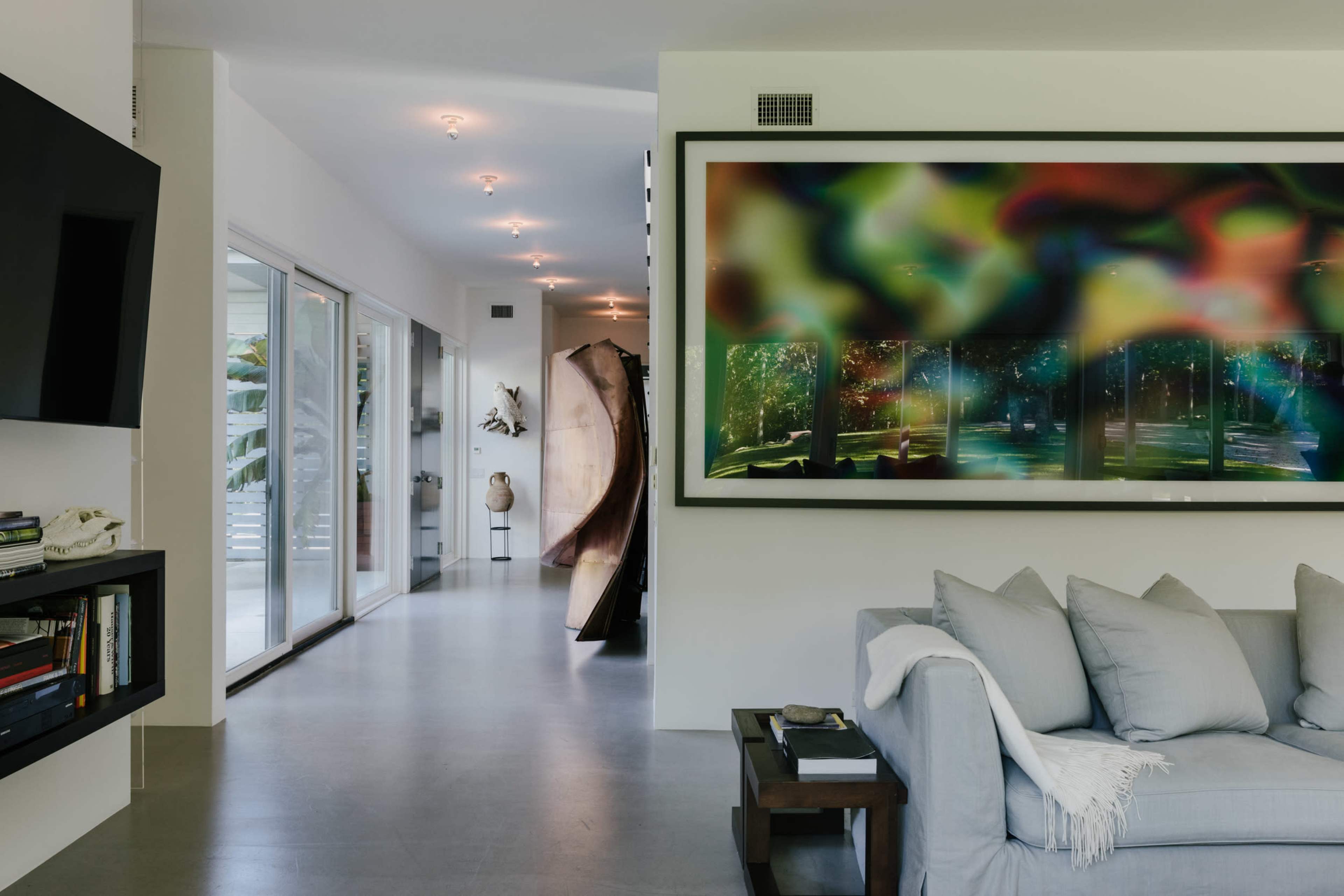 A modern interior space featuring a living room with a light gray sofa, a large colorful artwork on the wall, and a view into a hallway with sliding glass doors.
