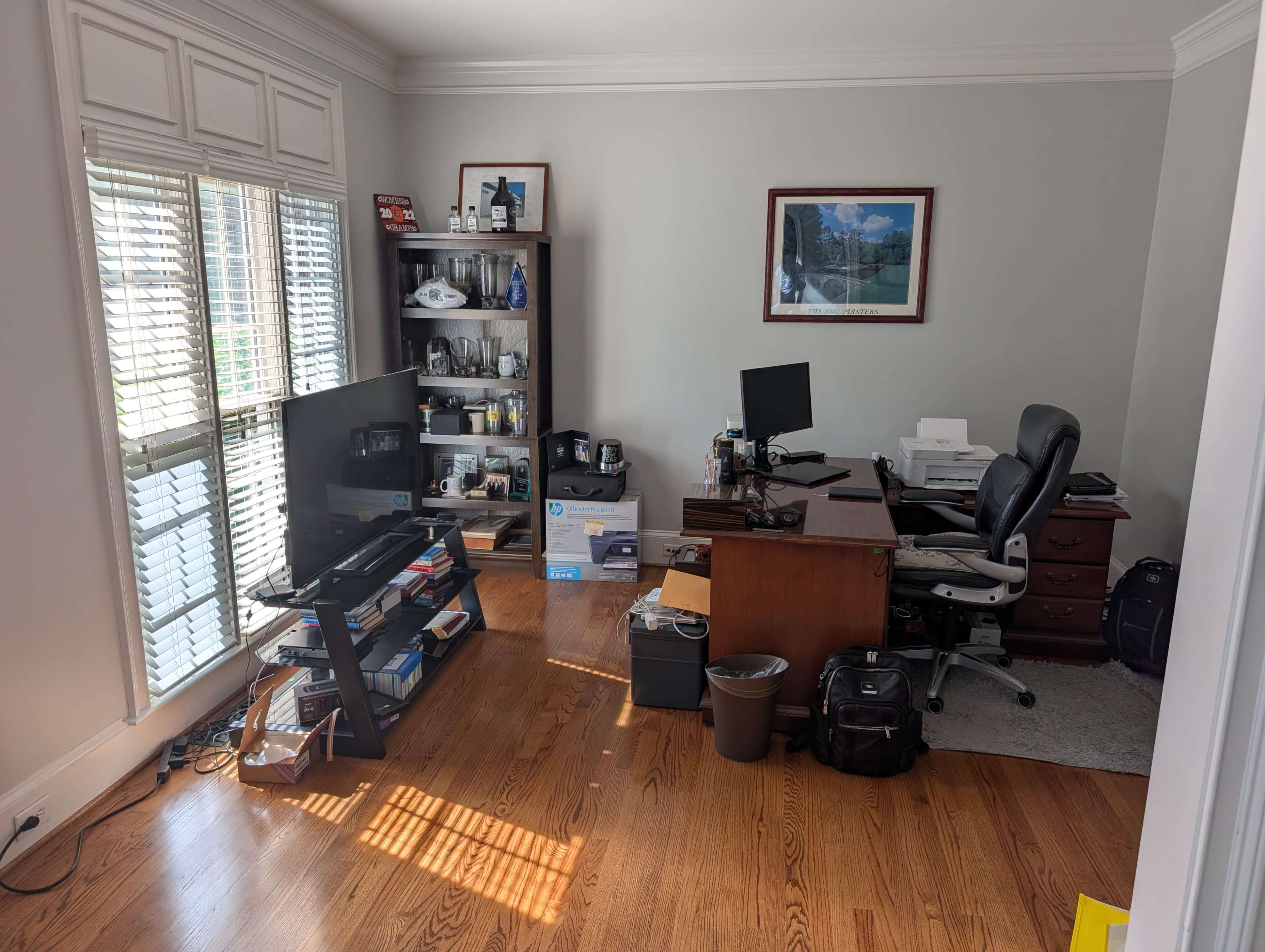 The image shows a home office with a wooden desk, two computer monitors, a bookcase filled with various items, a television, and a window with blinds, all situated on a wooden floor.