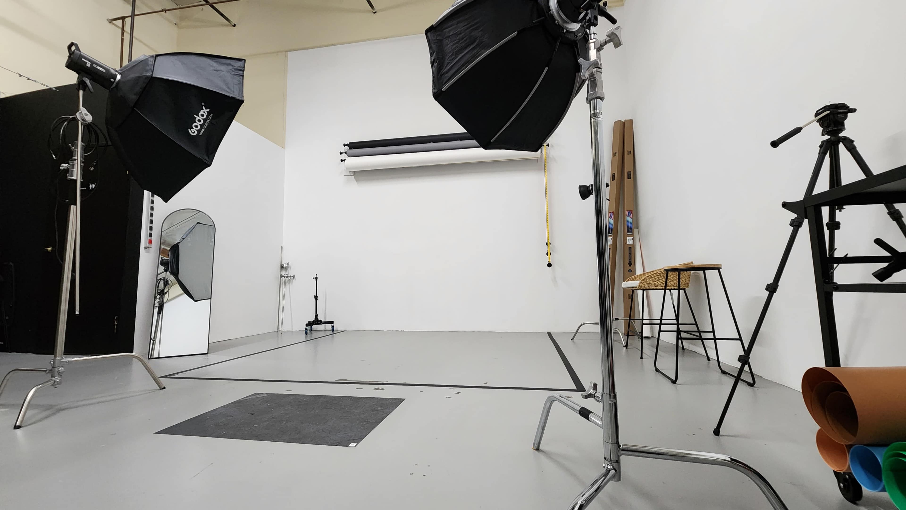 A photography studio with lighting equipment, a mirror, a stool, and rolls of backdrops against a white wall.
