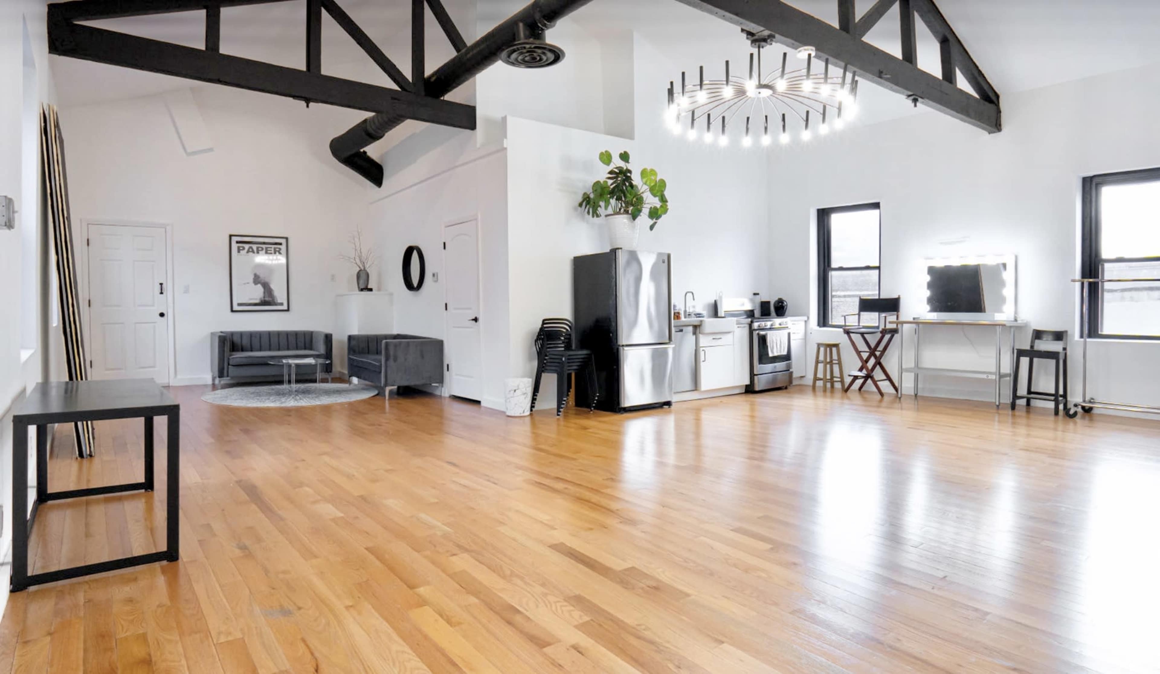 The image shows a spacious, modern interior with wooden floors, a kitchen area, and large windows, illuminated by a circular chandelier.
