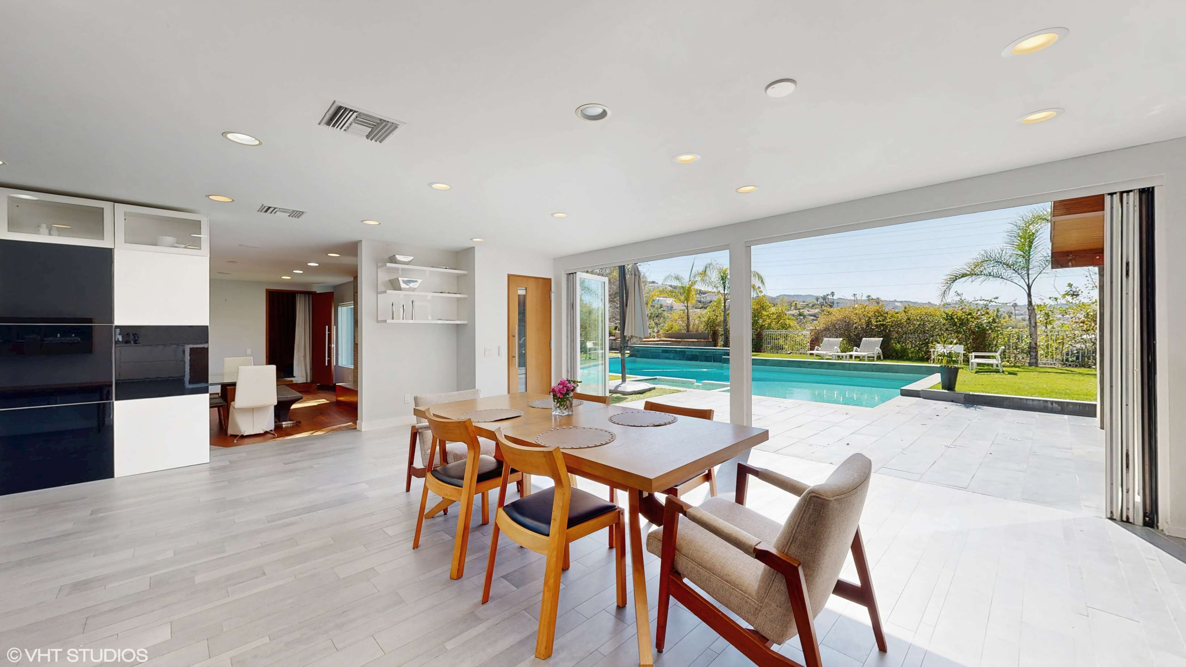 A dining area with a wooden table and chairs overlooks a backyard with a swimming pool and greenery.