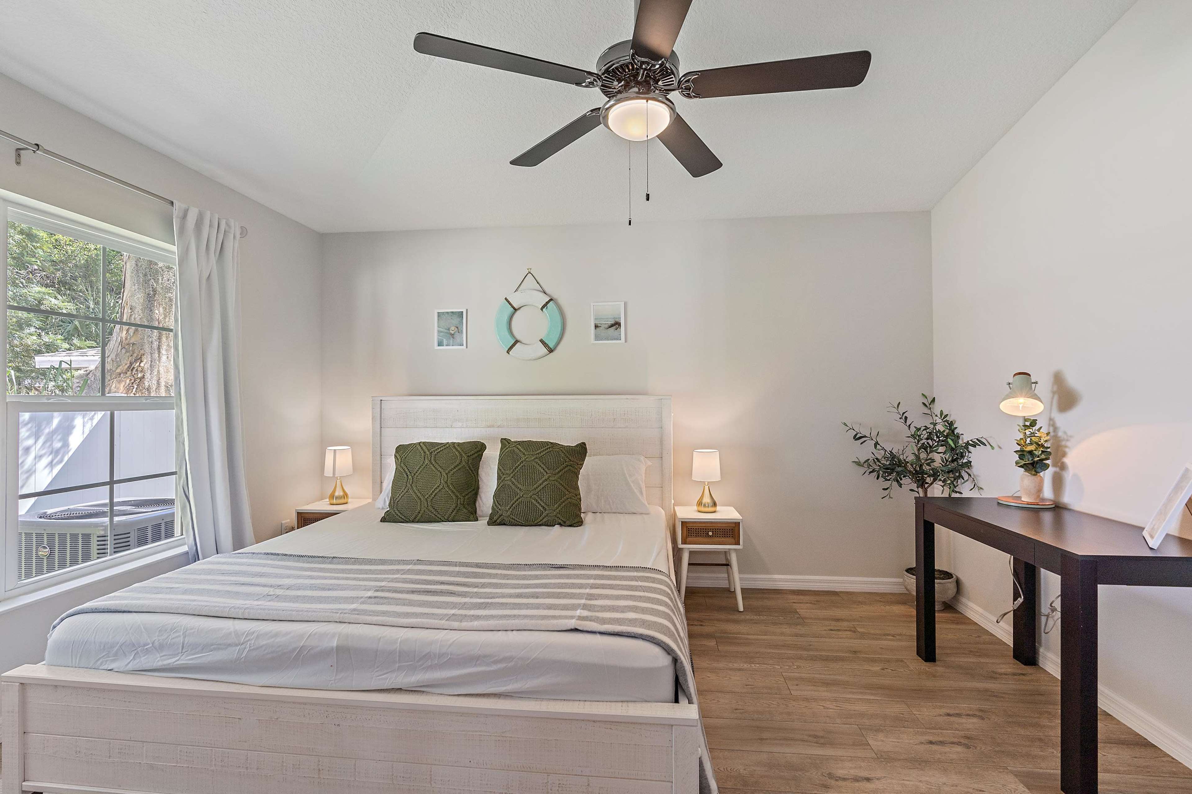 A well-lit bedroom features a queen-sized bed with white linens, two green decorative pillows, a wooden nightstand, and a desk, all arranged against light gray walls.