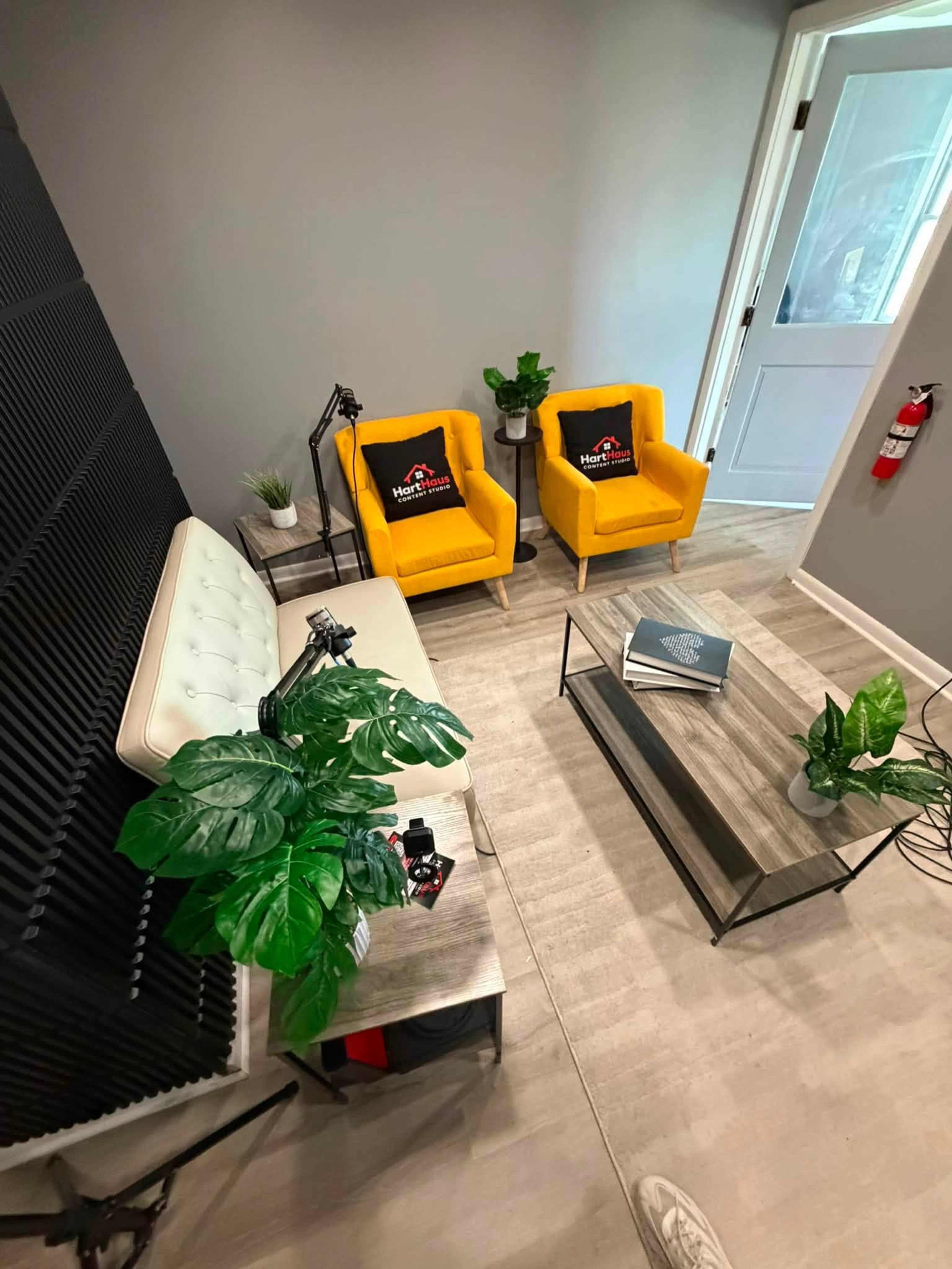 The image shows a modern small seating area with two yellow armchairs, a white sofa, a coffee table, and decorative plants, all set in a room with gray walls and a door leading to another space.