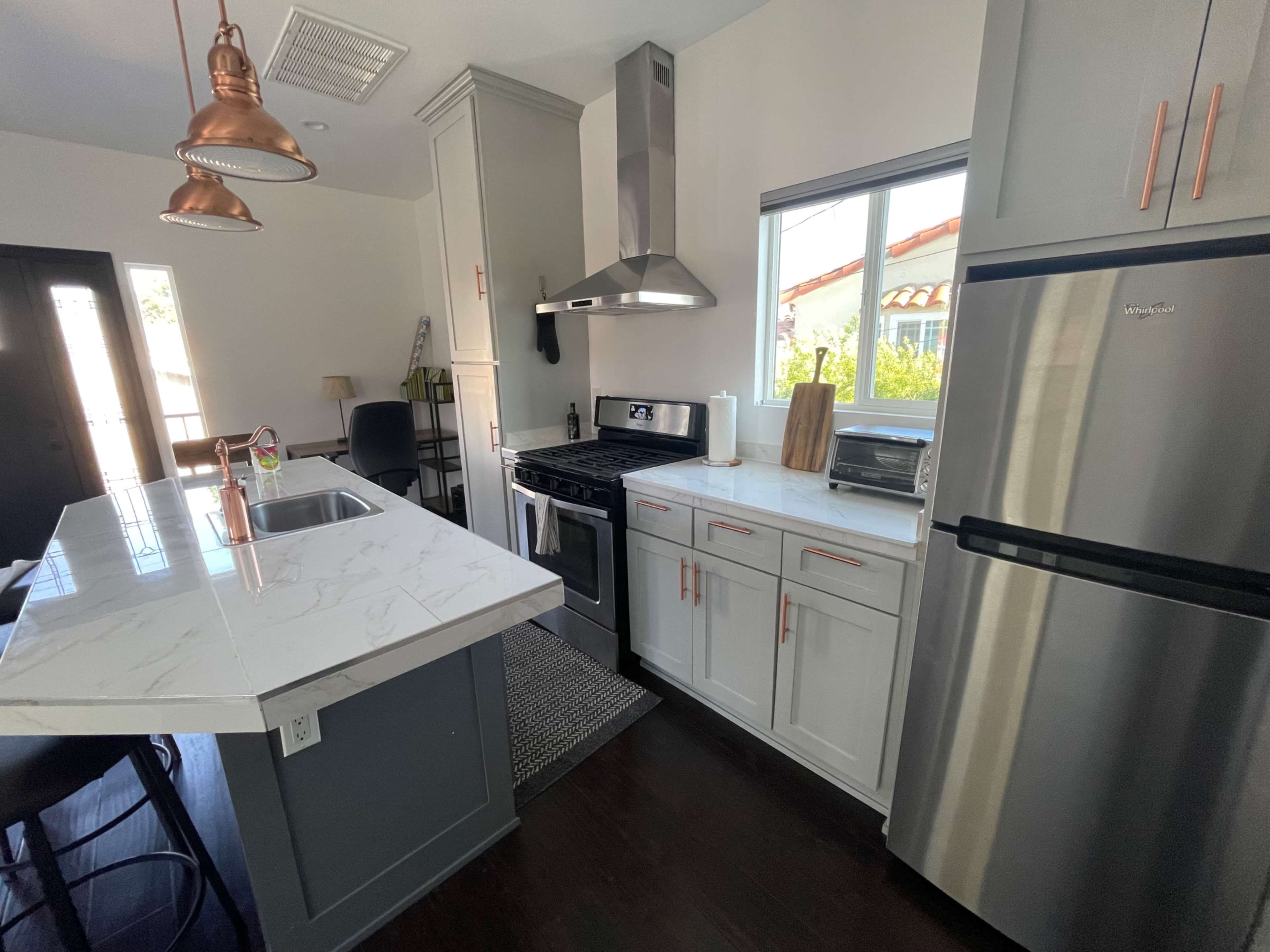 The image shows a modern kitchen featuring stainless steel appliances, a marble countertop island, and light gray cabinetry with copper accents.