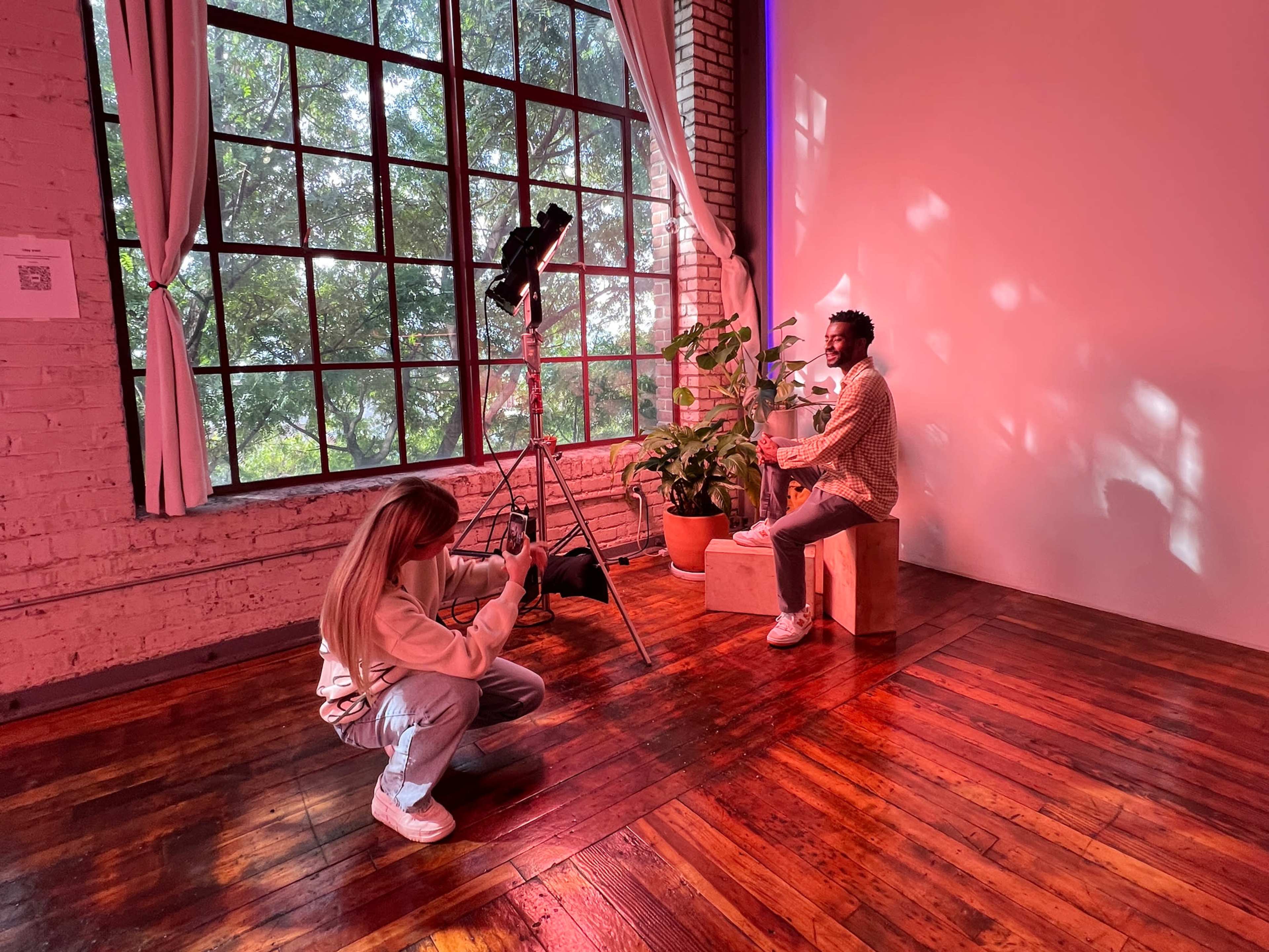 A woman operates a camera while a man poses on a wooden box in front of a large window adorned with greenery.