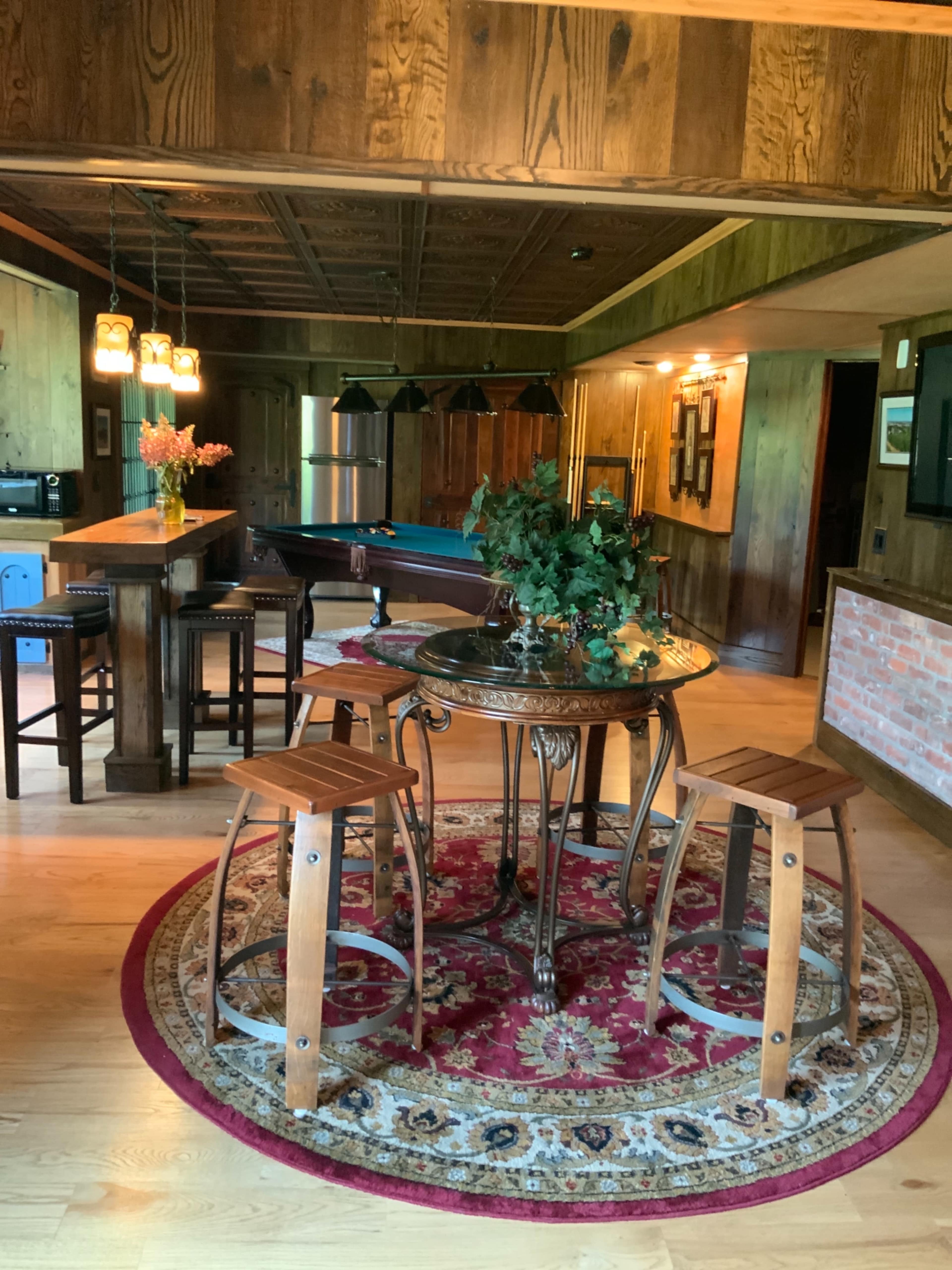 The image shows a rustic room with a round table surrounded by two wooden stools on a decorative rug, adjacent to a bar area and a pool table in the background.