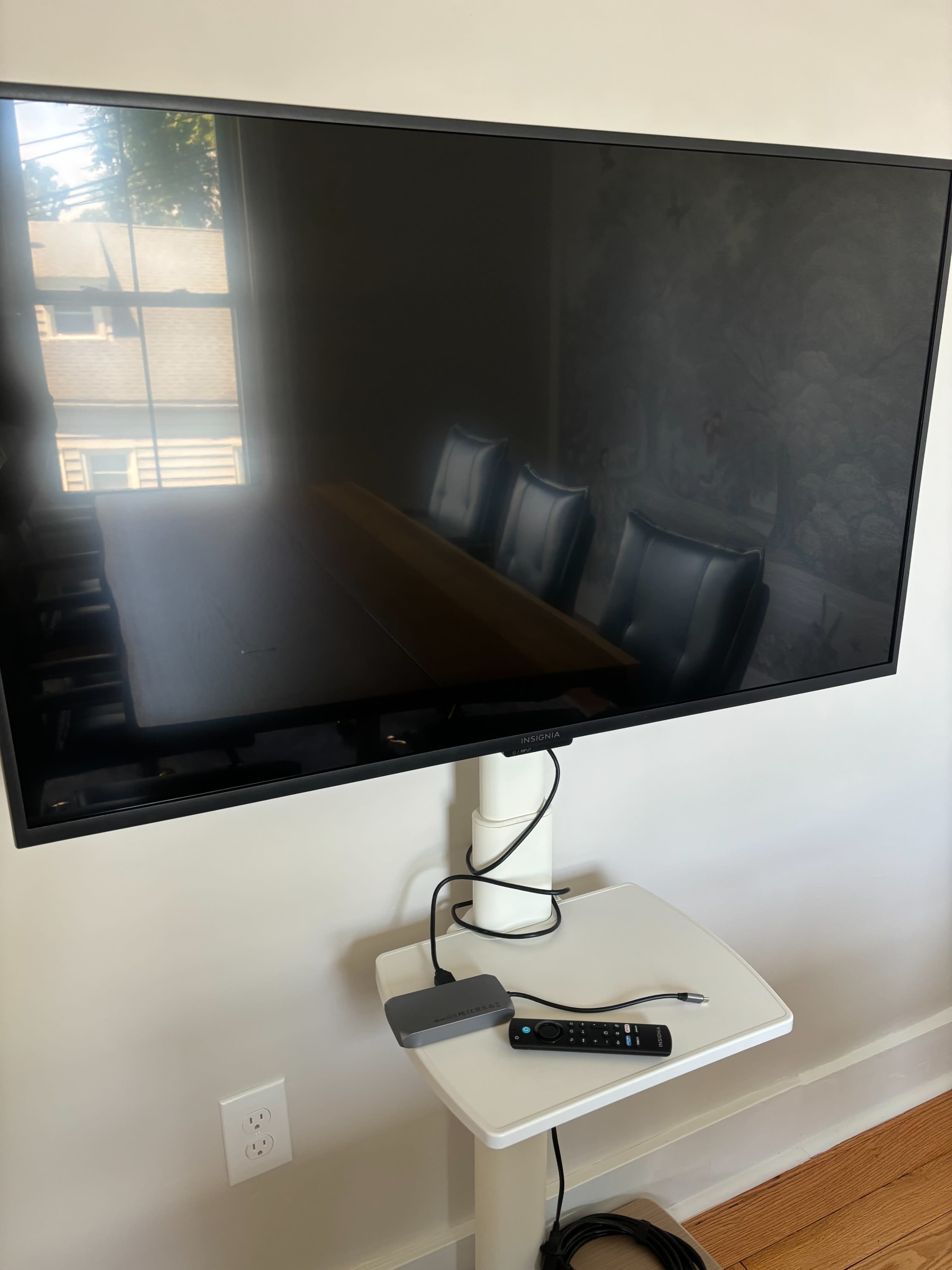 A large, unlit television is mounted on a white stand beside a wooden conference table with black chairs in a well-lit room.