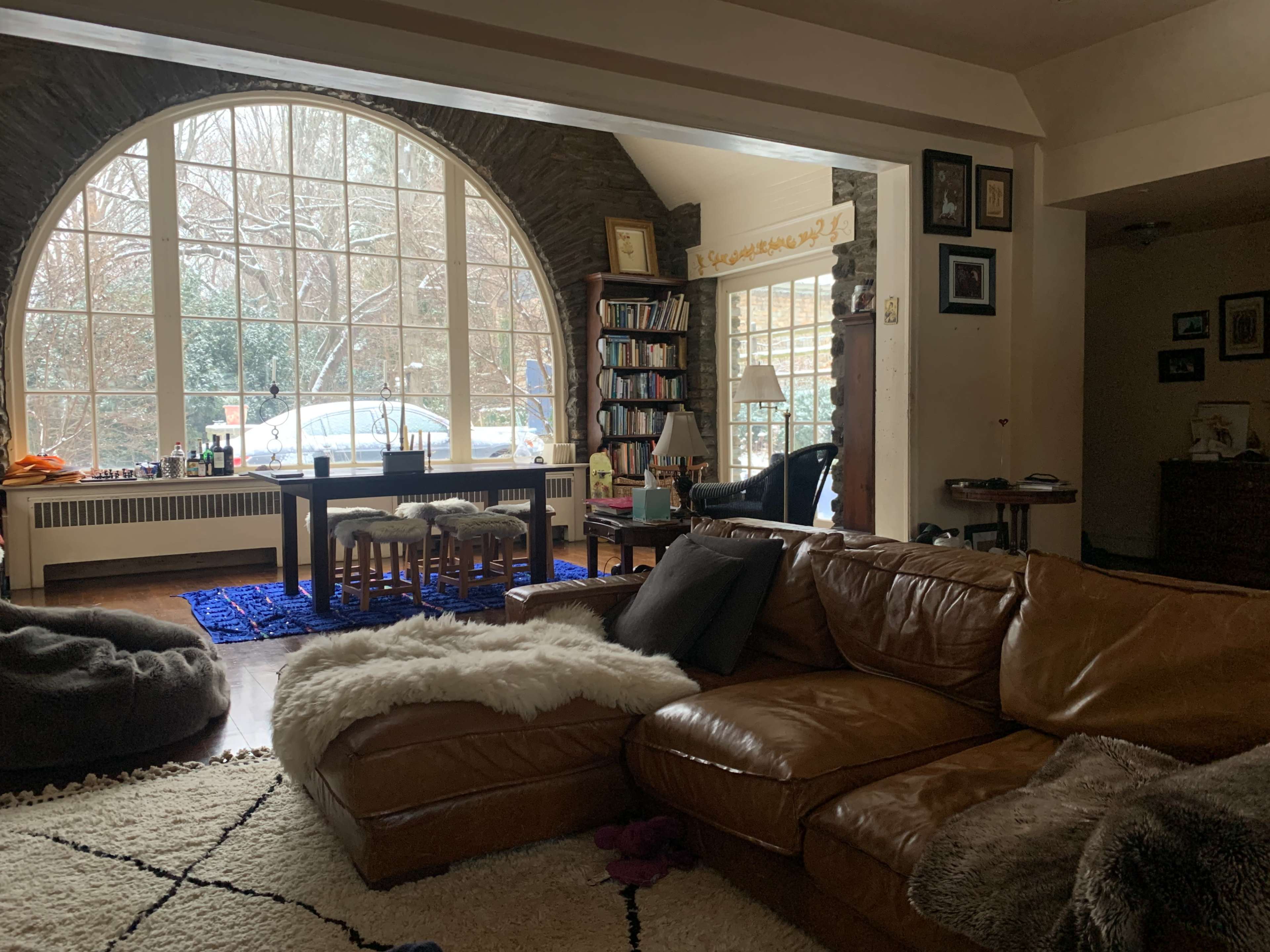 The room features a large arched window, a brown leather sectional couch, and a table with chairs surrounded by bookshelves.