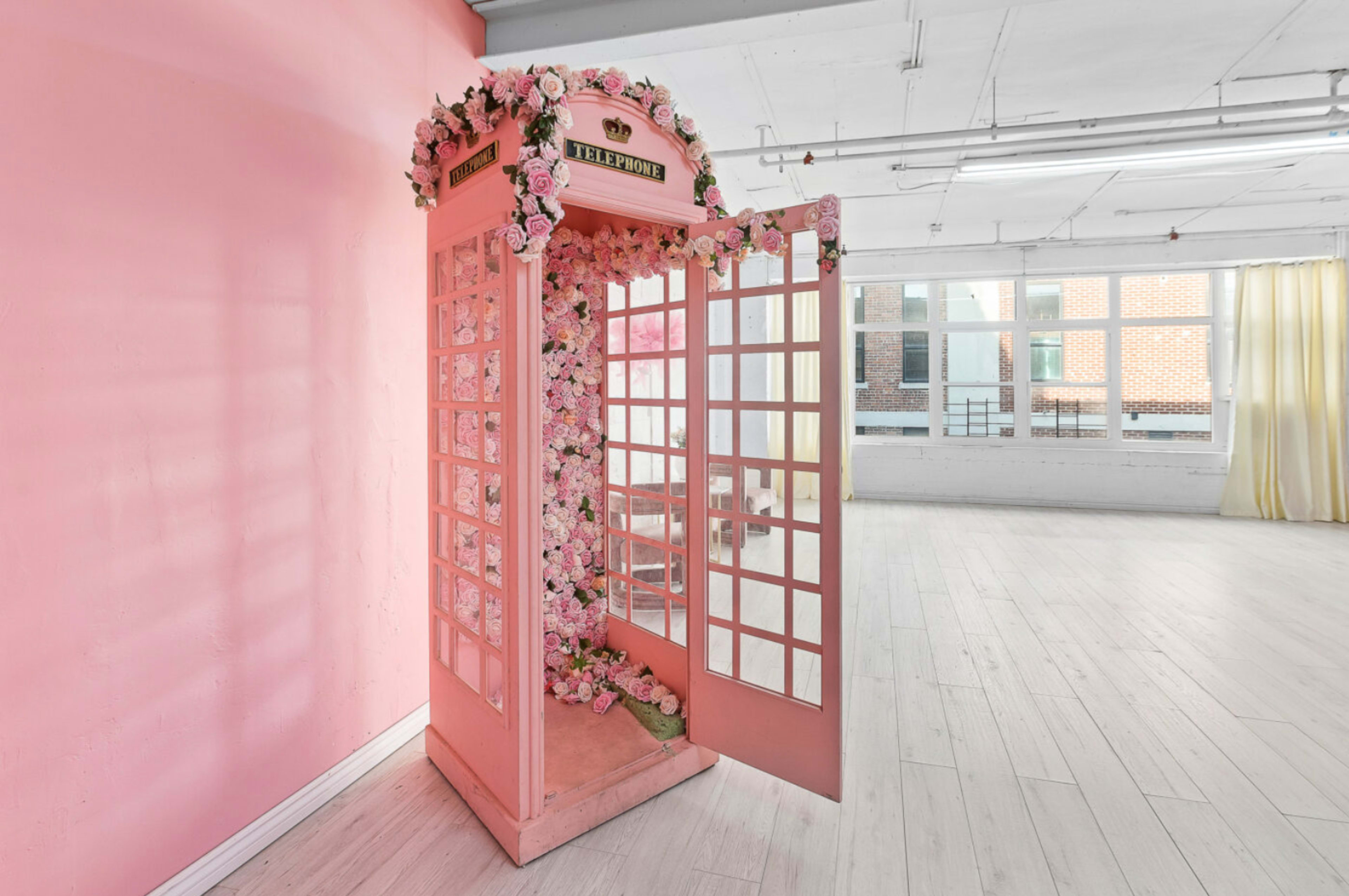 A pink telephone booth adorned with flowers stands in a bright, airy room with large windows.