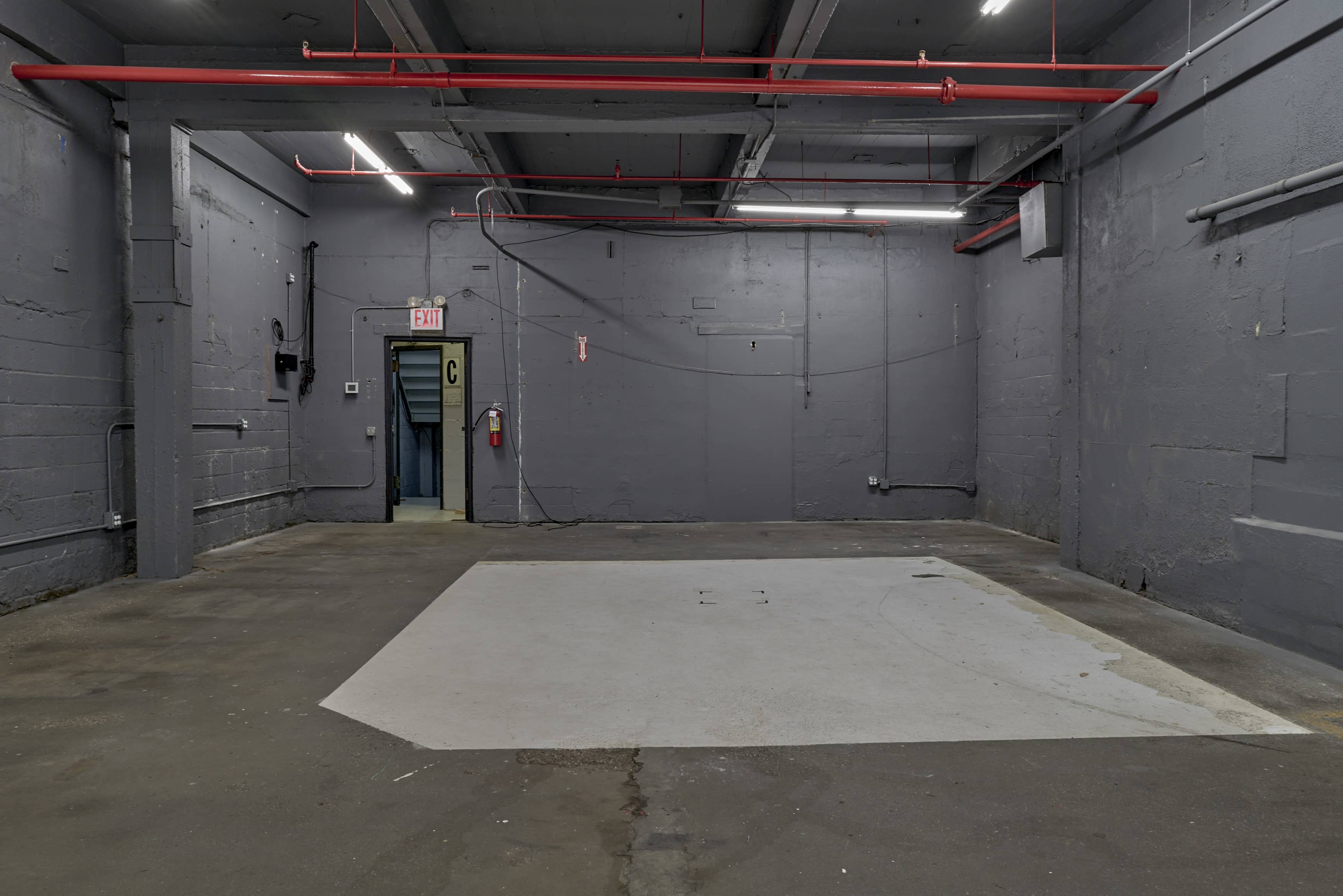 The image depicts an empty industrial space with gray walls and concrete flooring, featuring red pipes along the ceiling and a door marked "C" in the background.