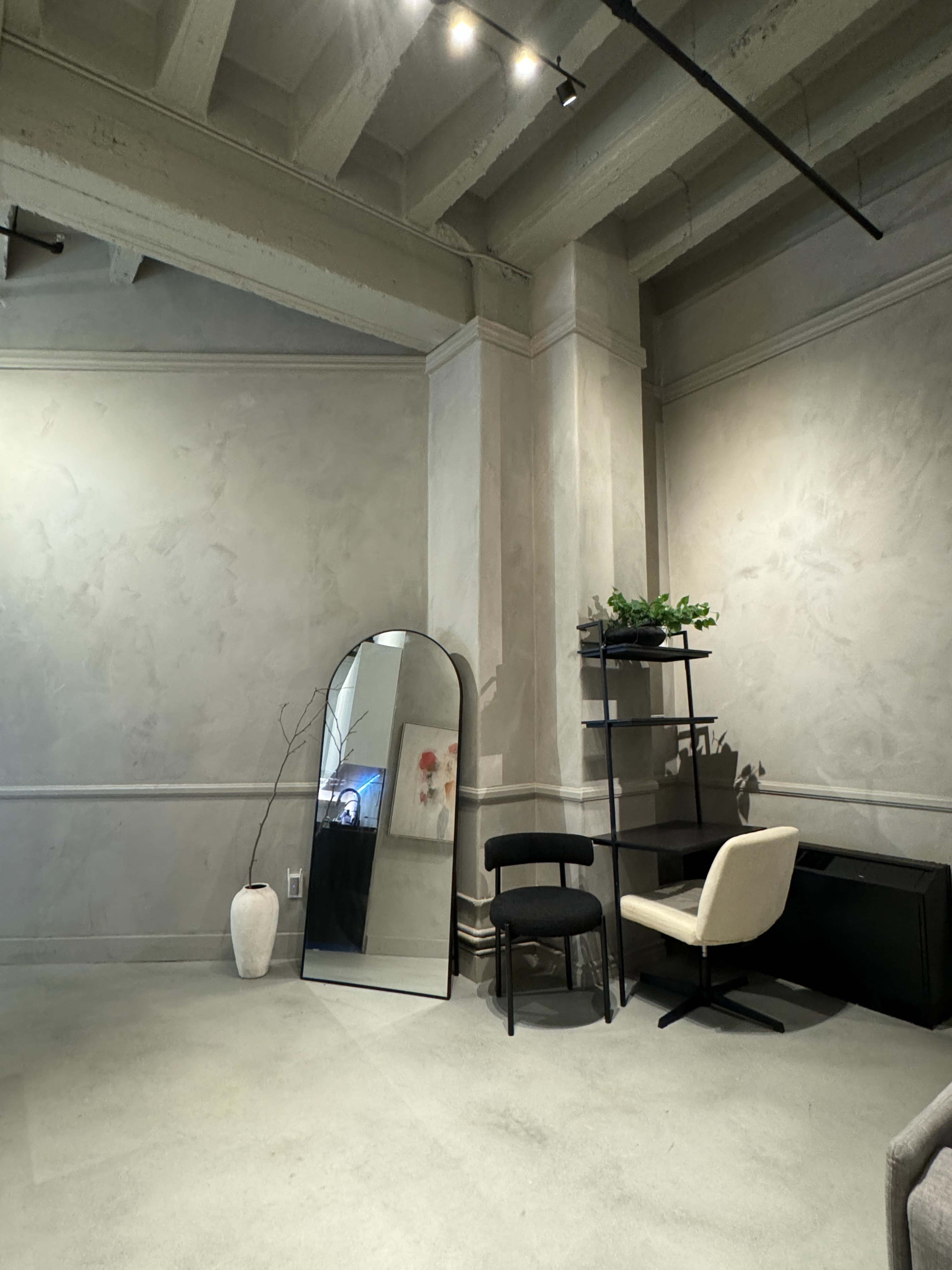 The image shows a minimalistic interior space featuring a curved mirror, a plant shelf, and two chairs against a textured wall.