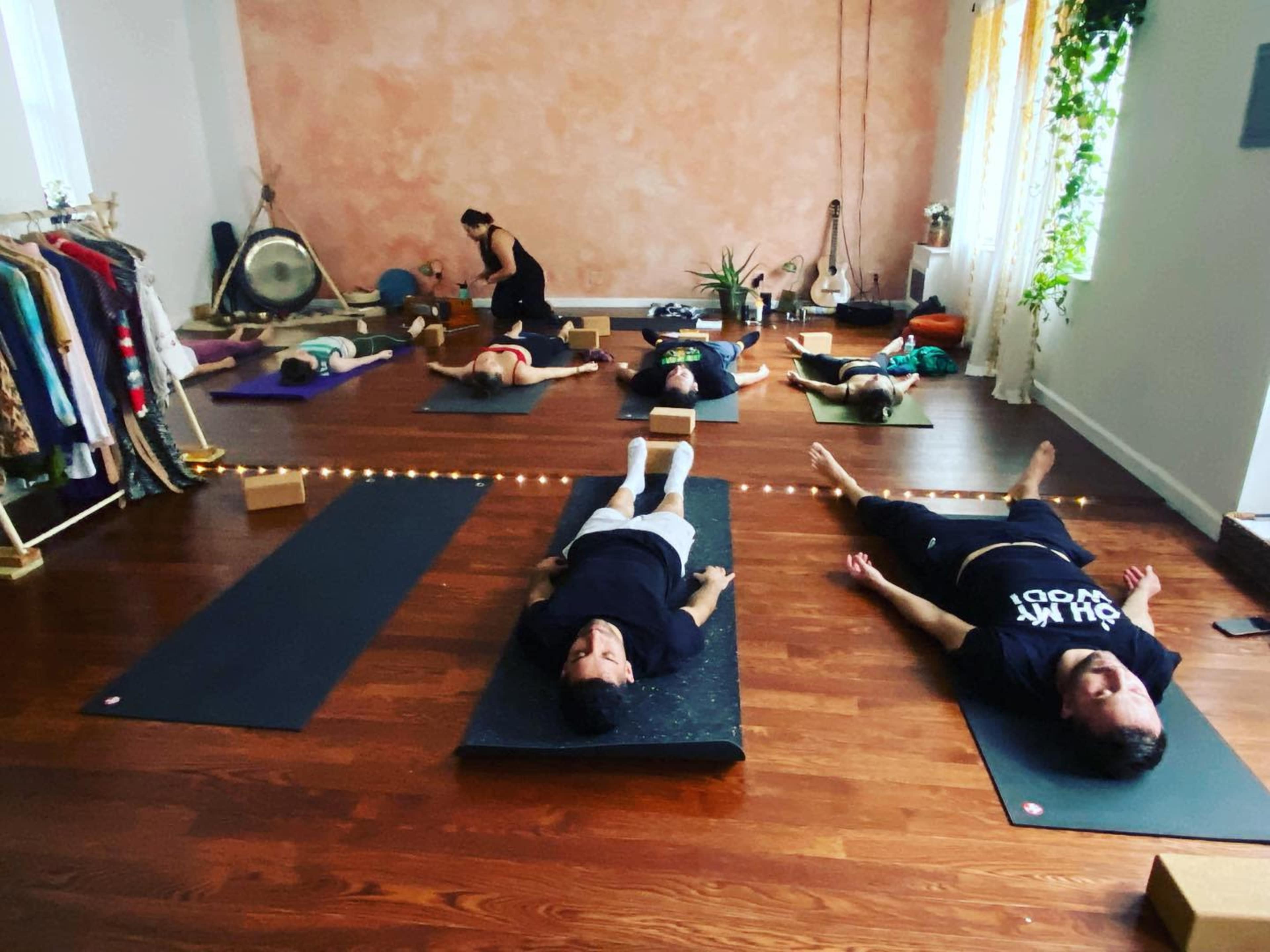 The image shows a yoga class in a studio with participants lying on mats, while a teacher leads at the front.