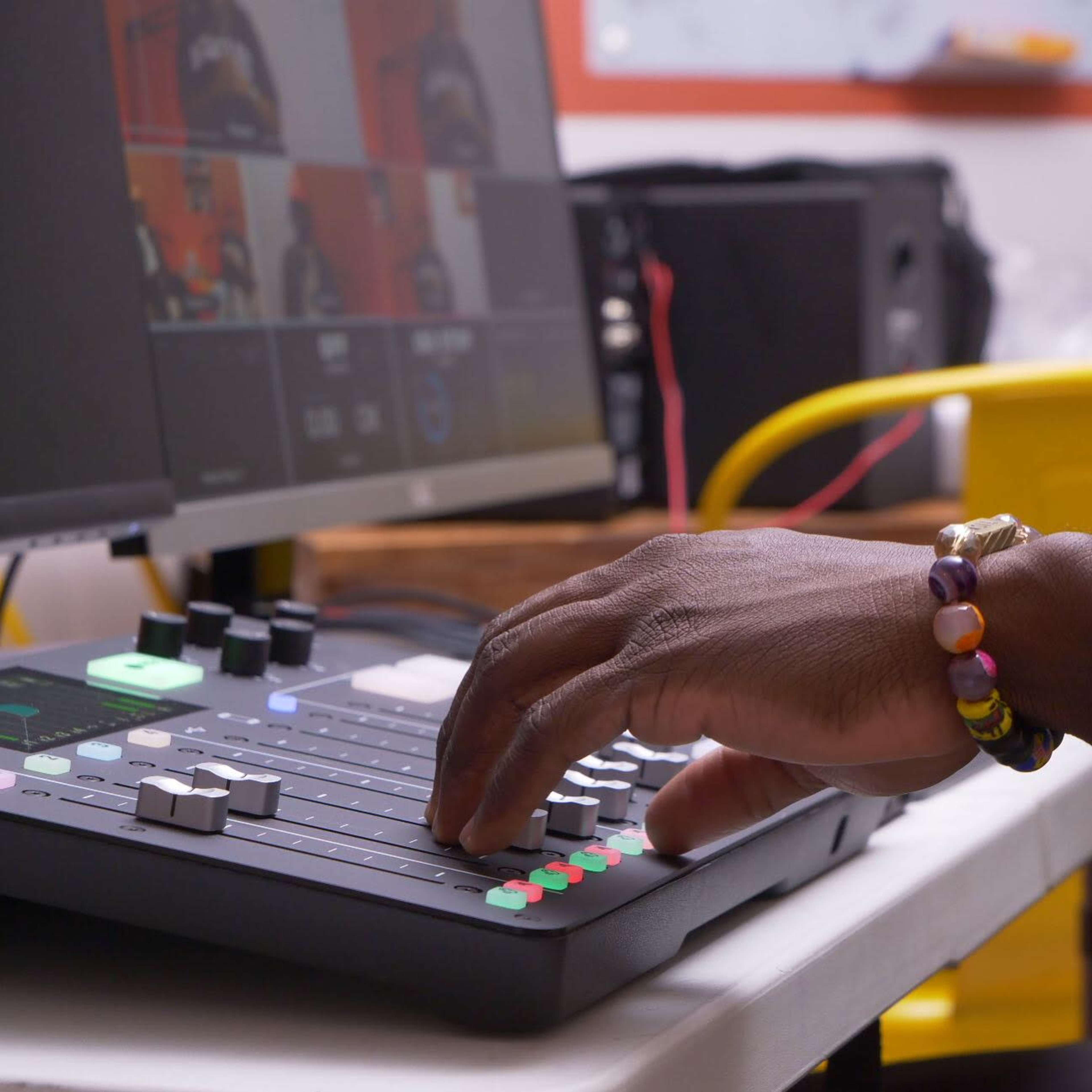 A hand adjusts the controls on a multimedia mixer next to a computer monitor displaying multiple video feeds.