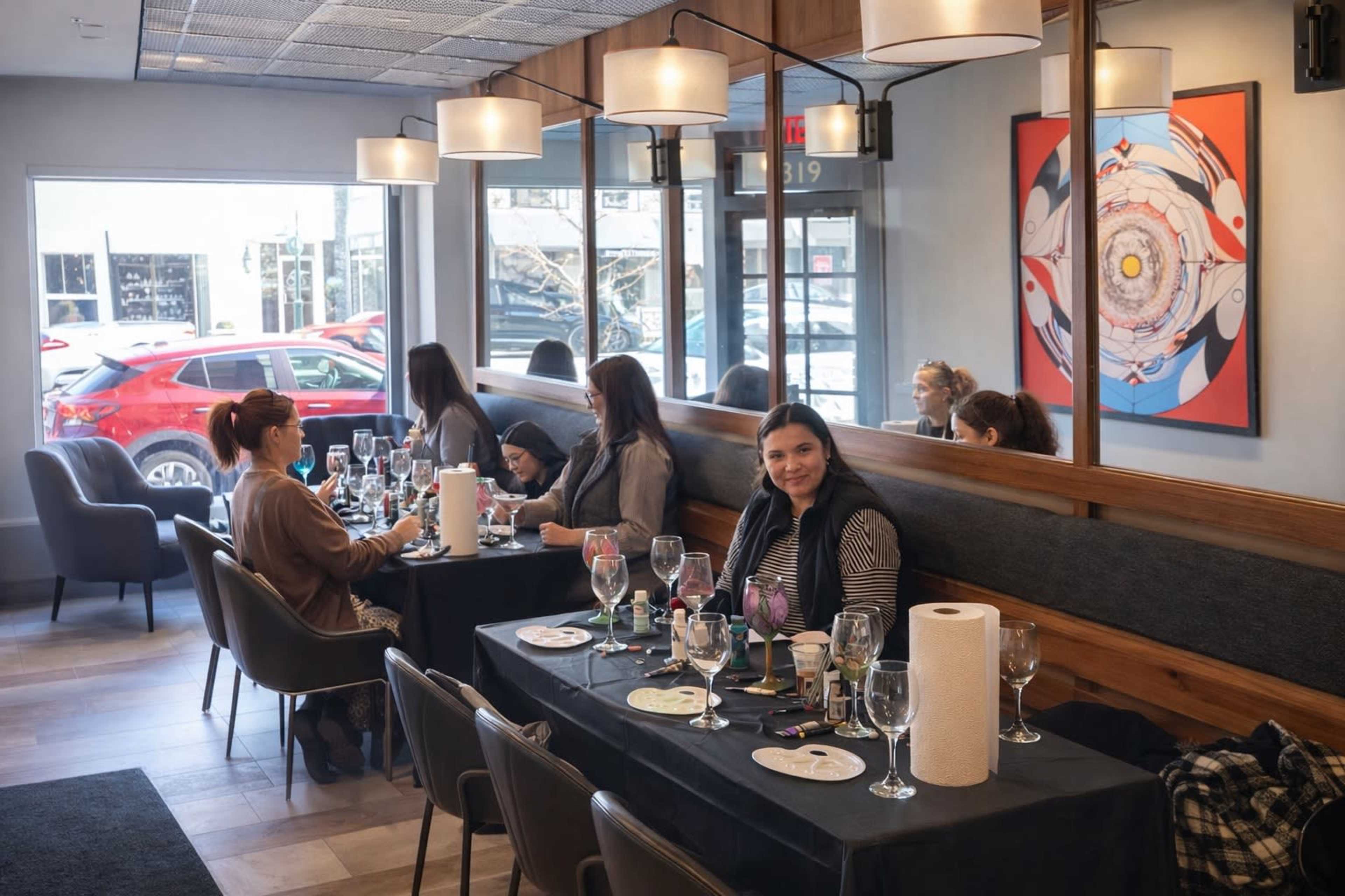The image shows a modern restaurant interior with several tables occupied by people, and a woman sitting at the foreground table with wine glasses and painting supplies.