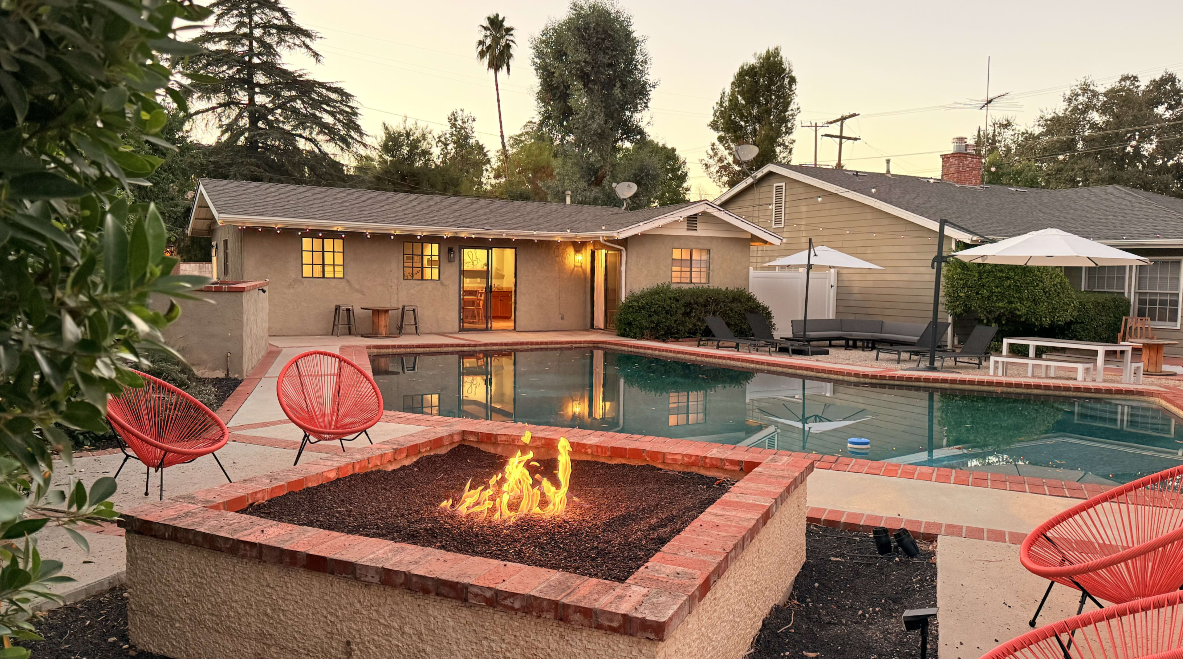 The scene depicts a backyard with a swimming pool, a fire pit with flames, and a cozy house in the background, illuminated by warm lights during dusk.