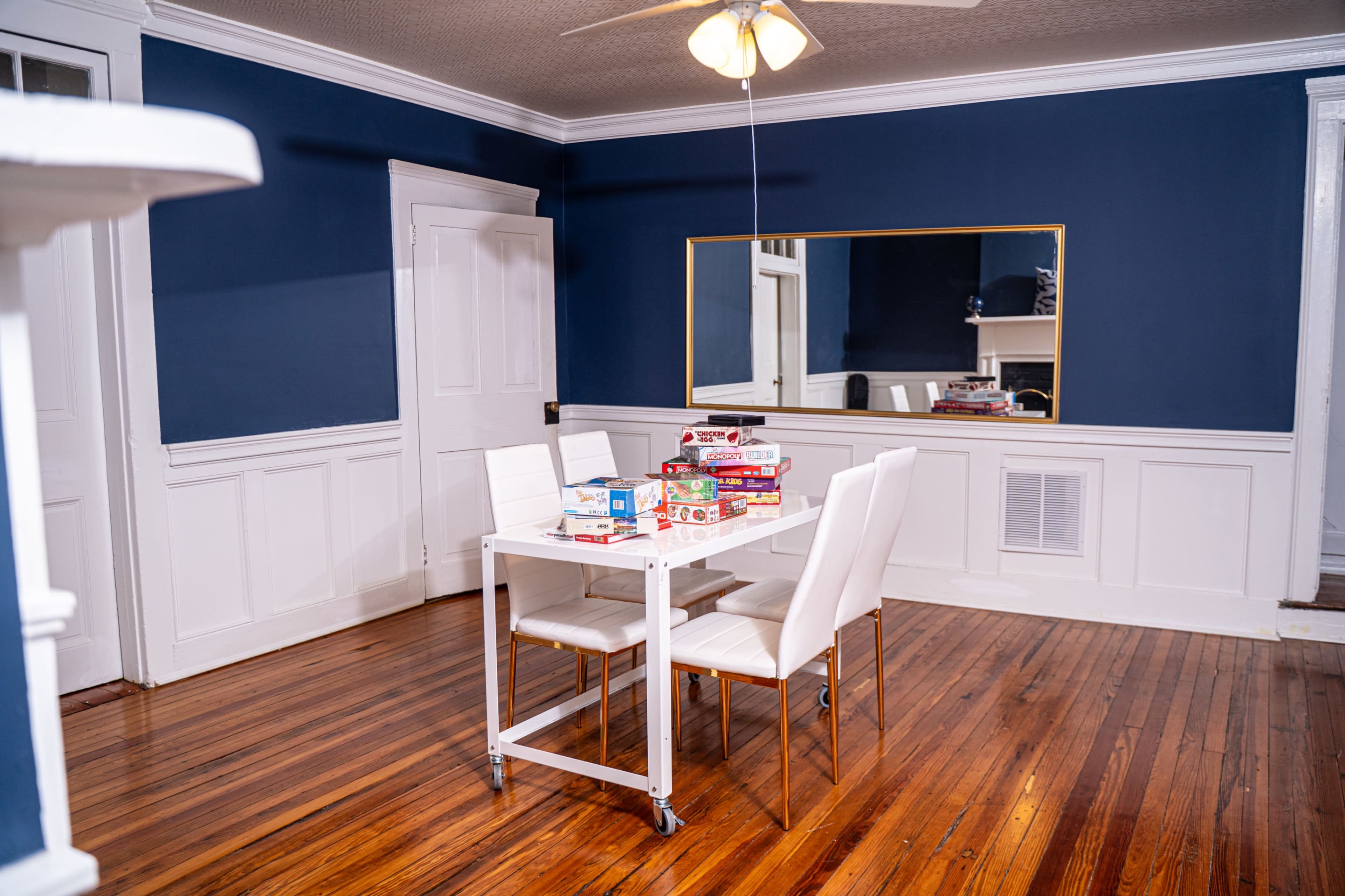 The image shows a dining area with a white table and four white chairs, a mirror on the wall, and a collection of board games stacked on the table.