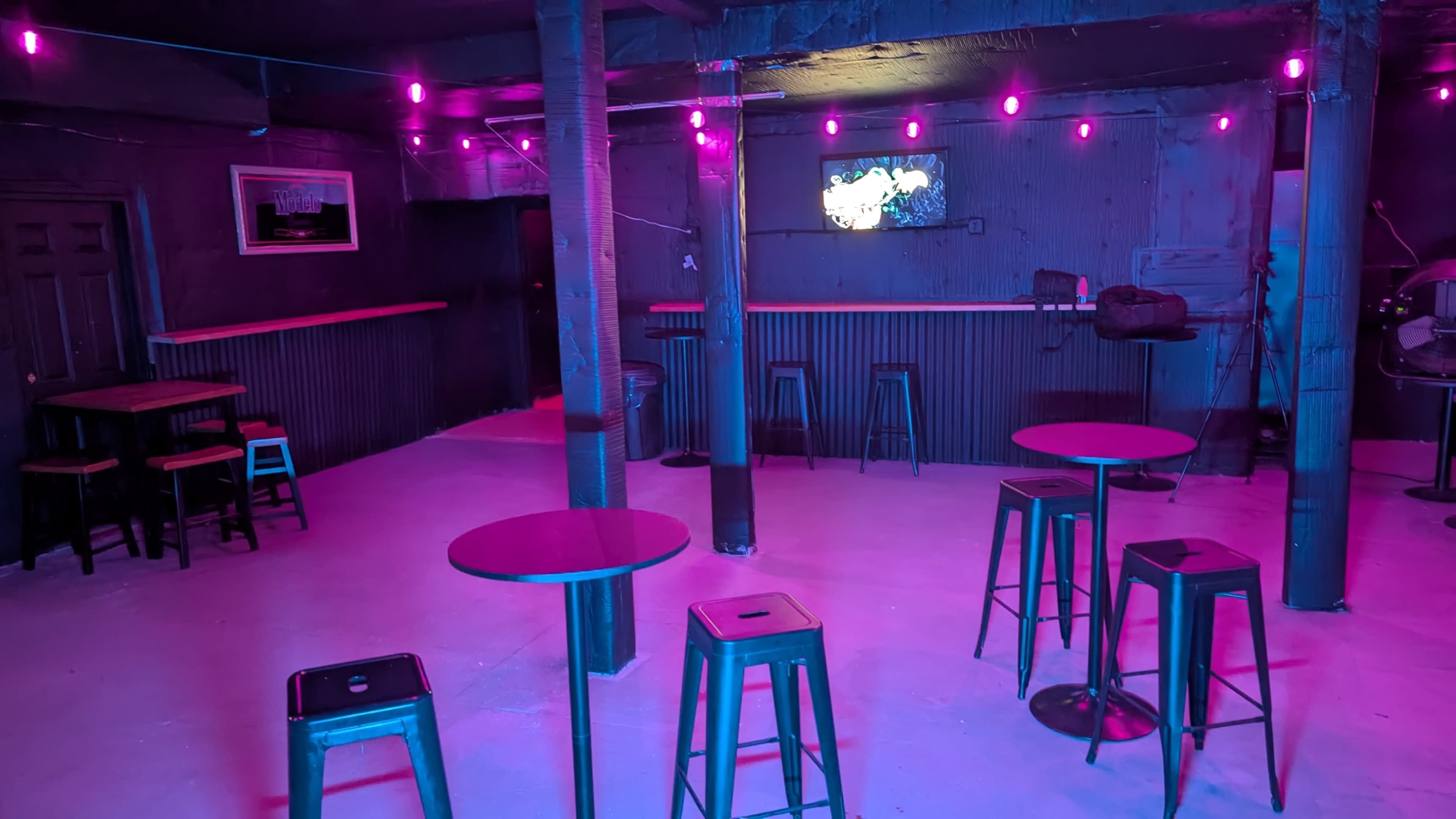 The interior of a dimly lit bar features several high-top tables and stools, with pink LED lights illuminating the space.