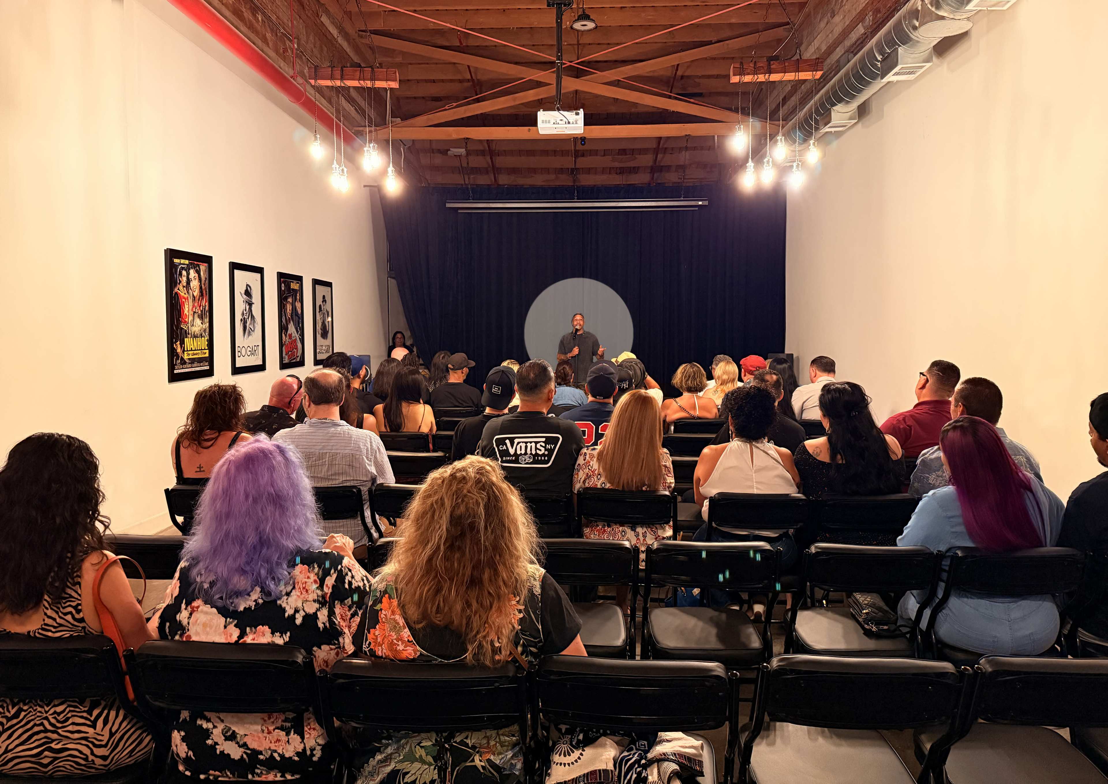 A speaker stands at the front of a dimly lit room filled with an audience seated in black chairs.