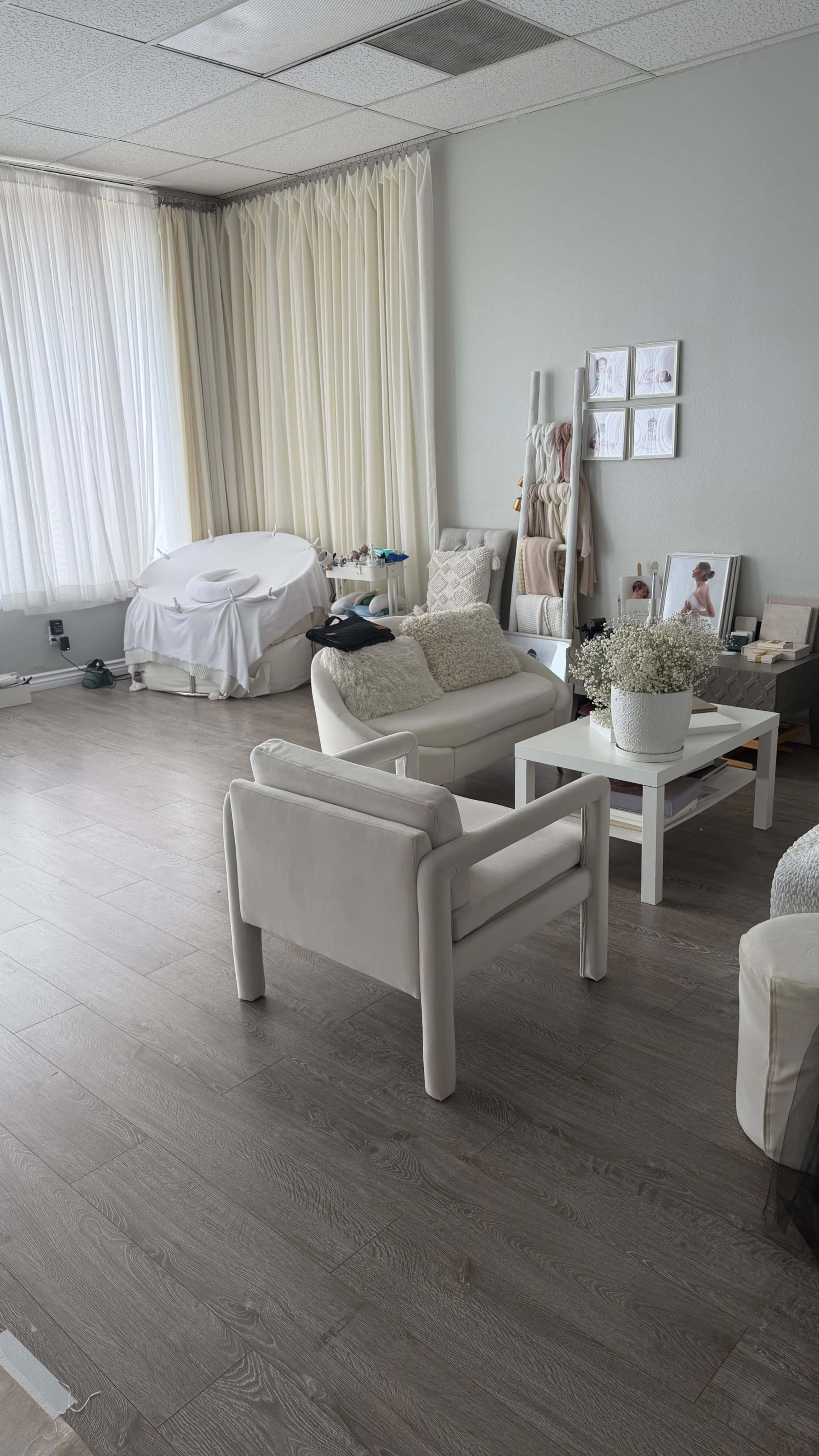 The image shows a bright, minimalist room with light-colored furniture, a large window with sheer curtains, and various decorative items arranged around the space.