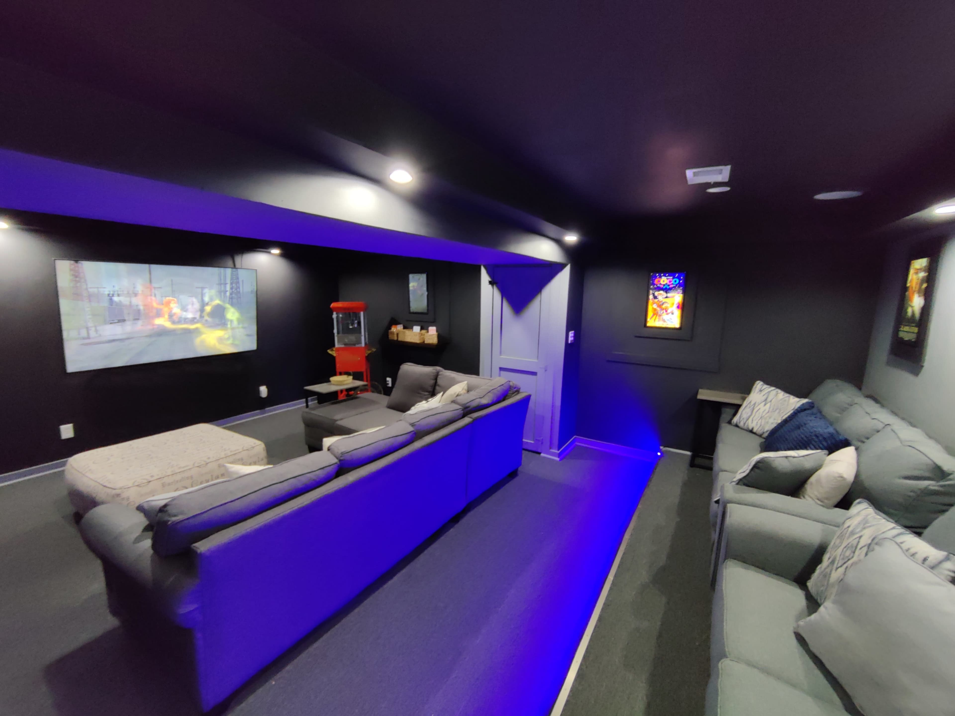 The image shows a home theater room with dark walls, comfortable seating, and a movie projected onto one wall along with a popcorn machine in the corner.