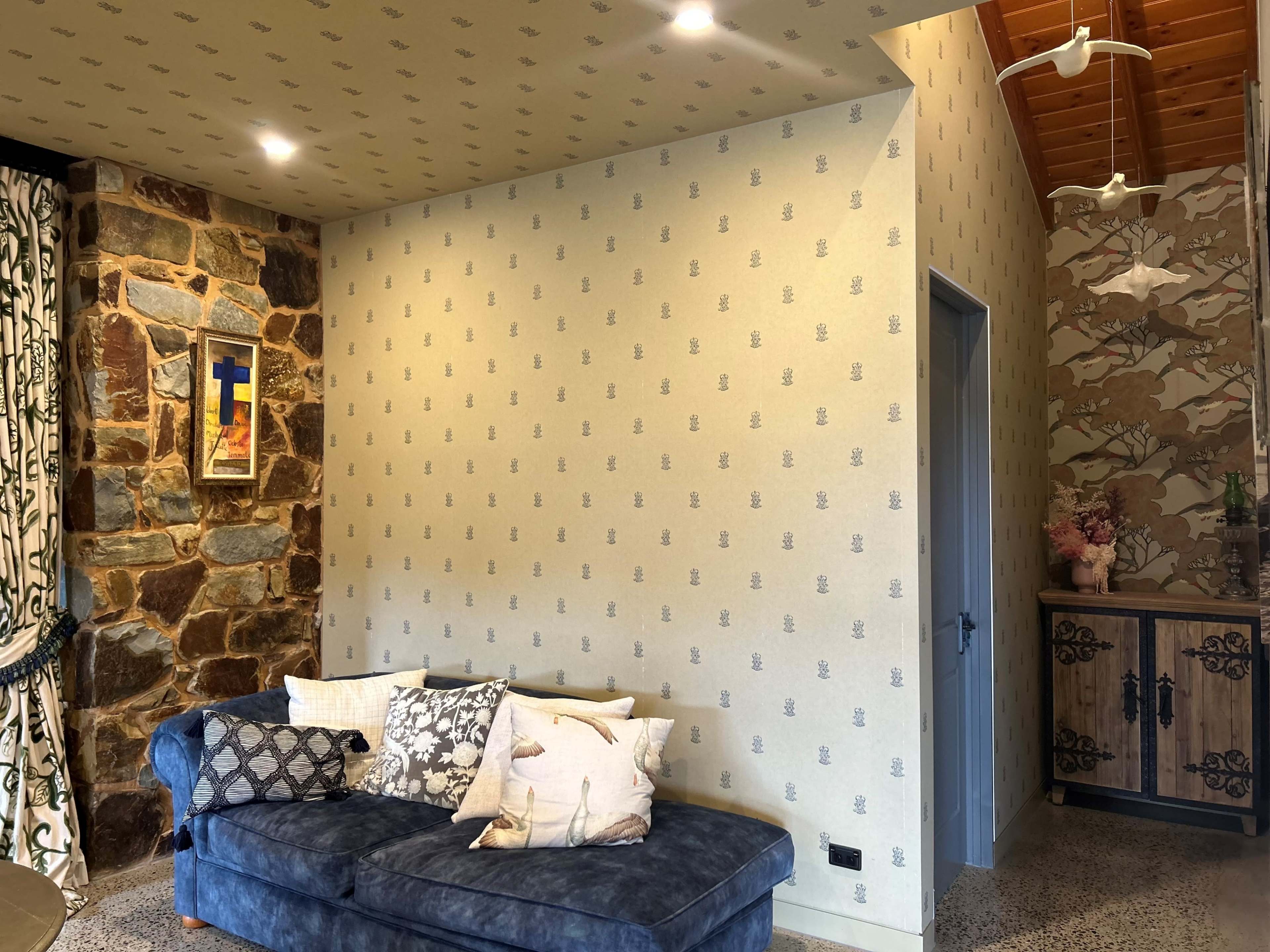 A living room corner featuring a blue sofa with decorative pillows, a patterned wall, and a doorway leading to another space.