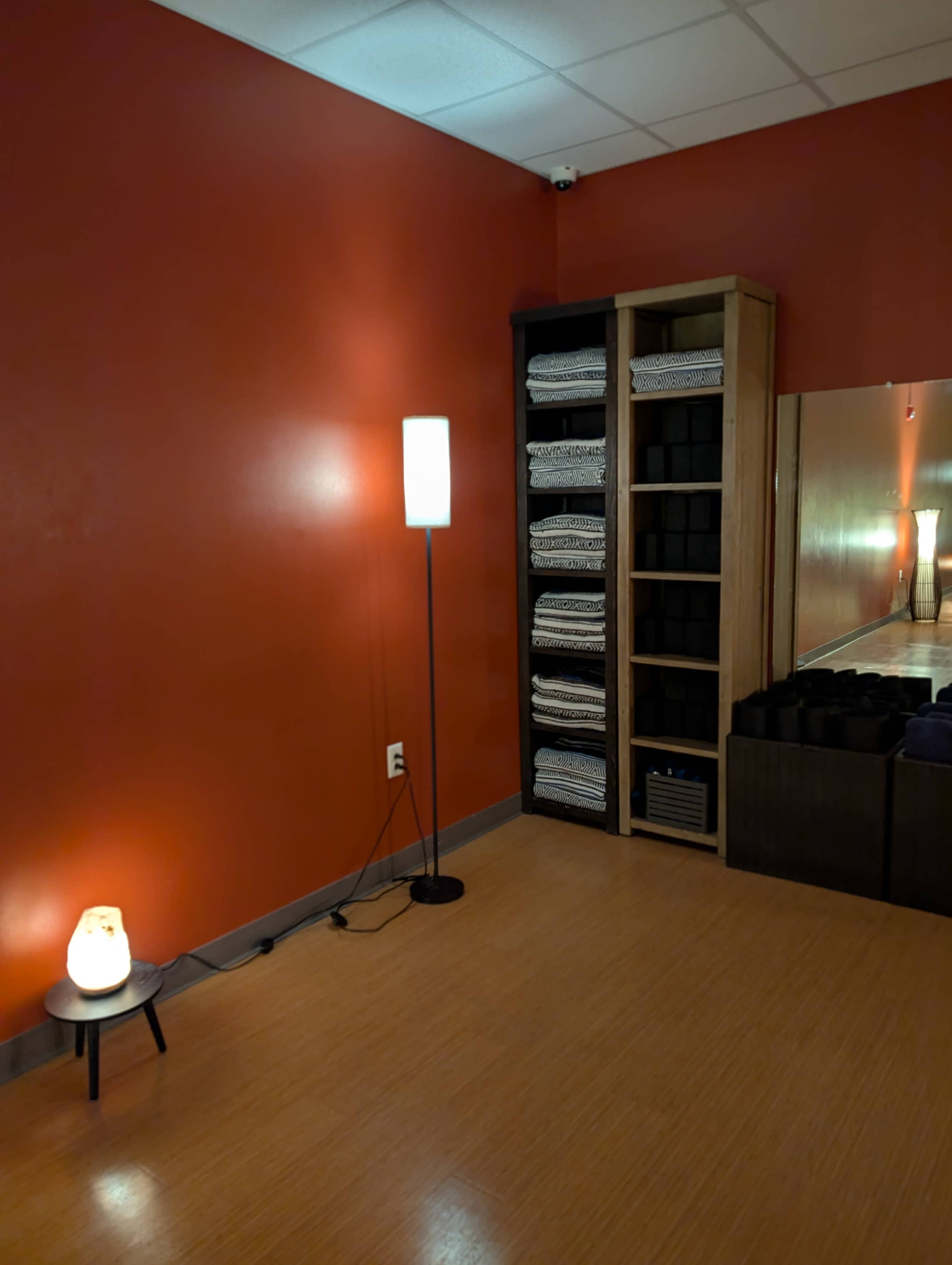 The image shows a calm, minimalistic room with red walls, a lamp, a shelf filled with neatly rolled towels, and a mirror reflecting the interior.
