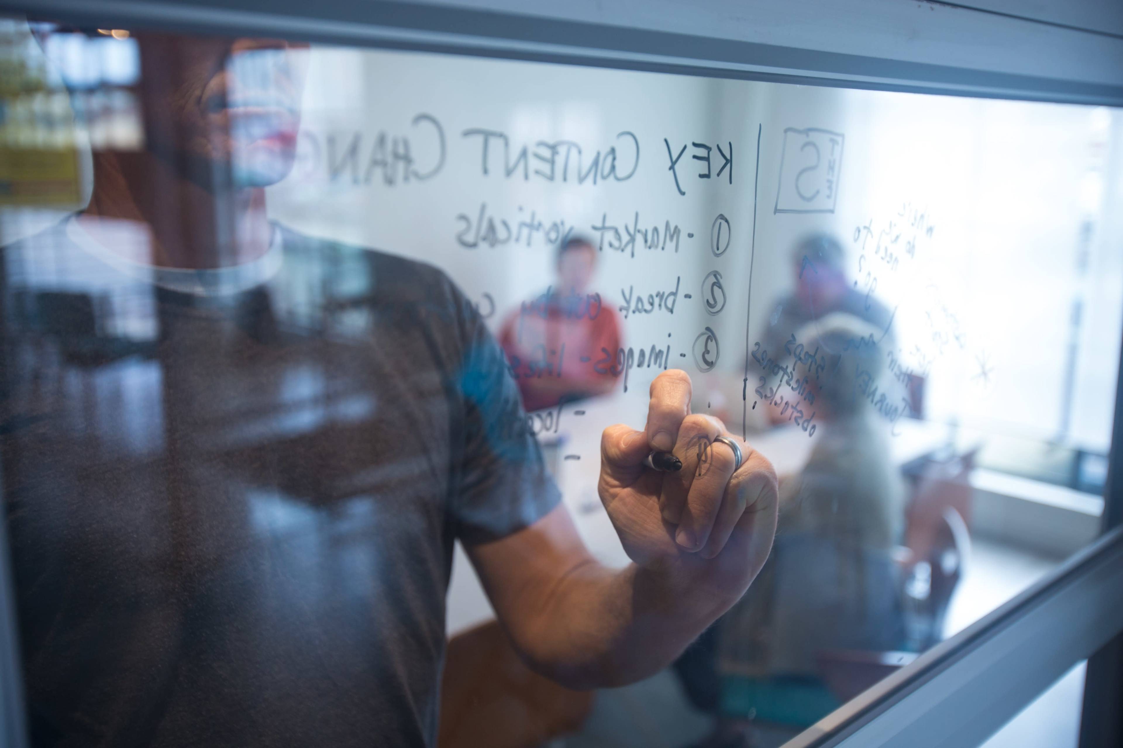 A person writes on a glass board while others sit at a table in the background.