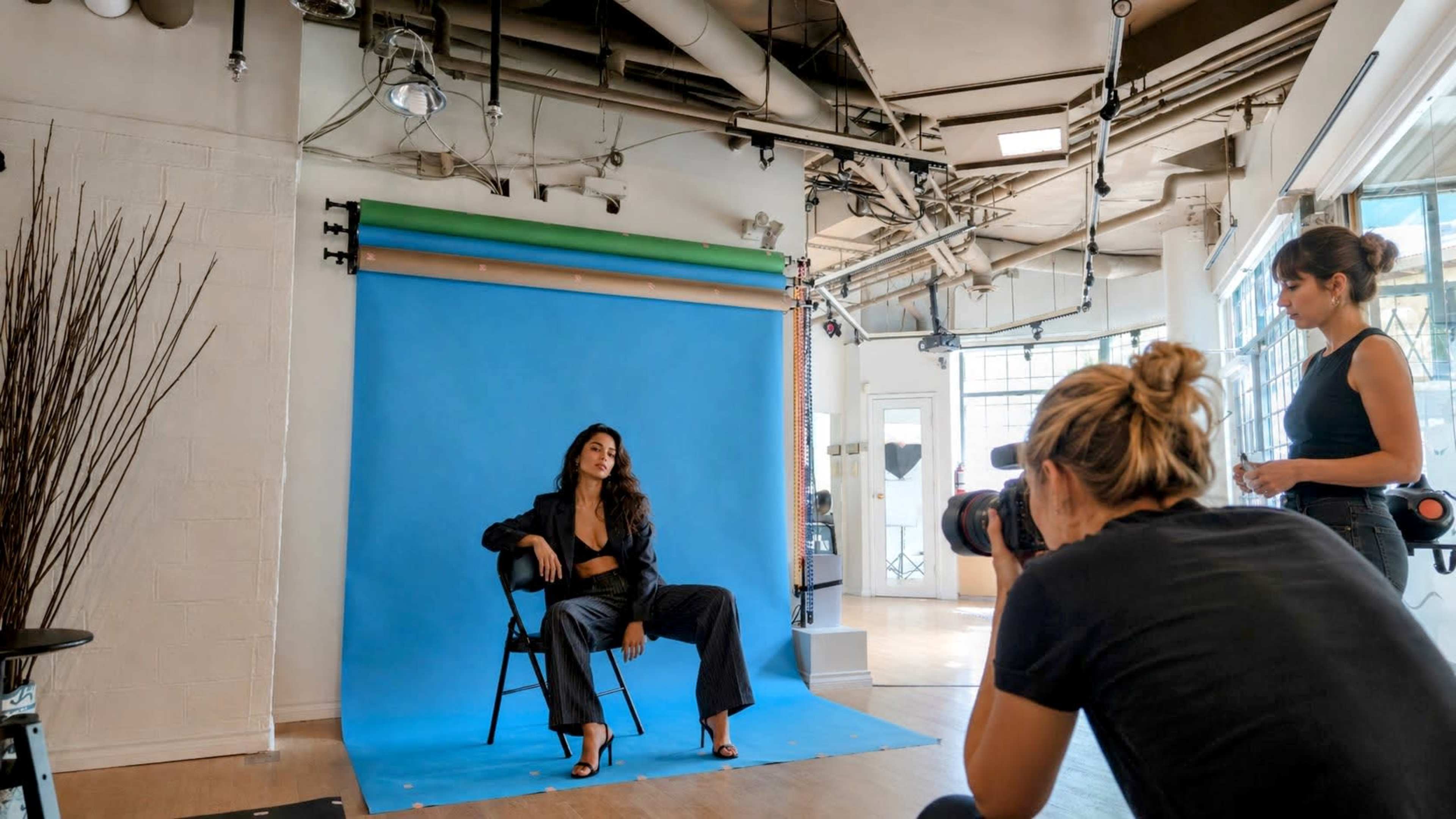 A model poses in a chair against a blue backdrop while a photographer captures the shot in a studio.