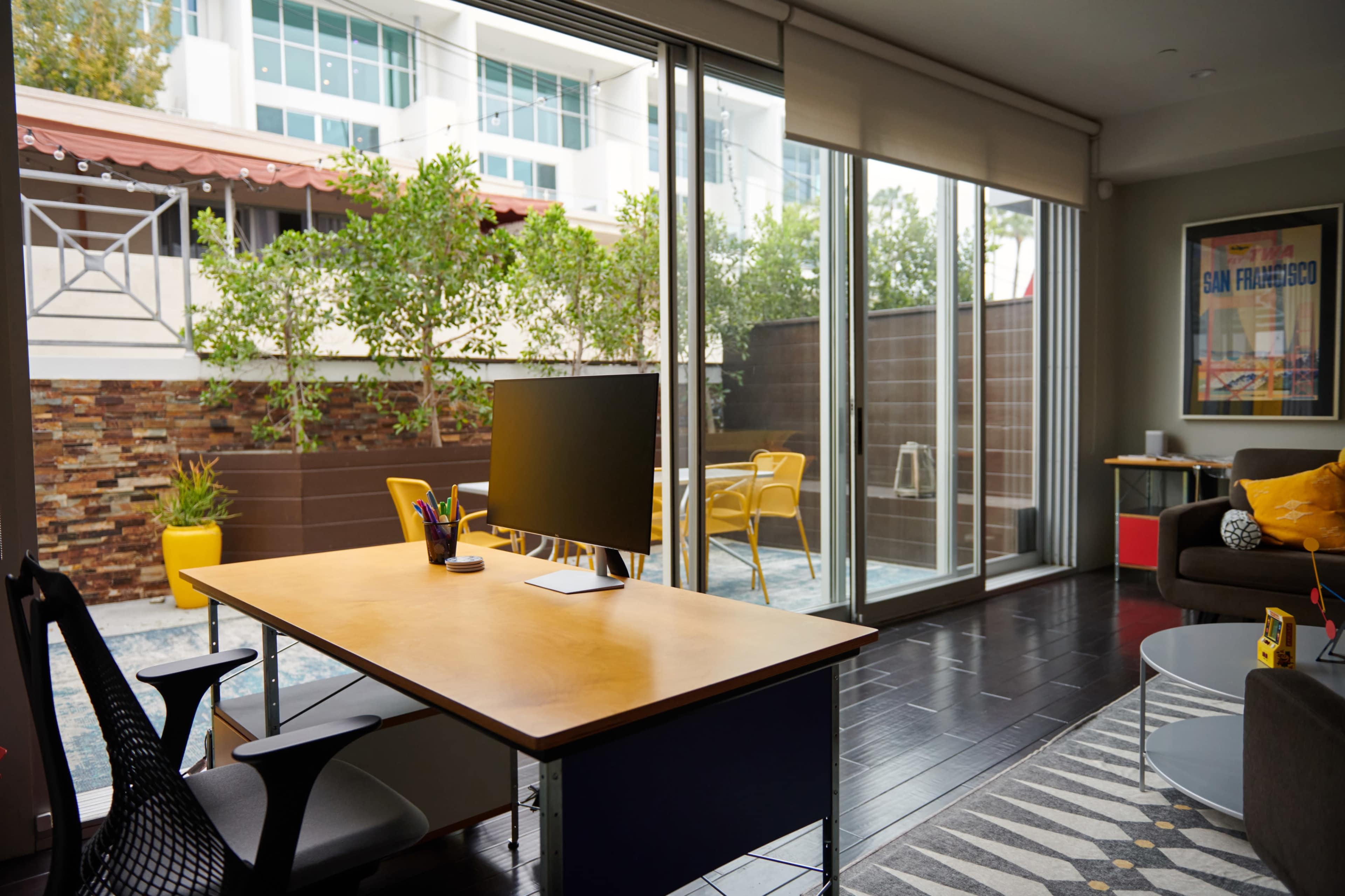 A modern home office with a desk and computer, facing a glass wall that opens to a small patio with yellow chairs and greenery outside.