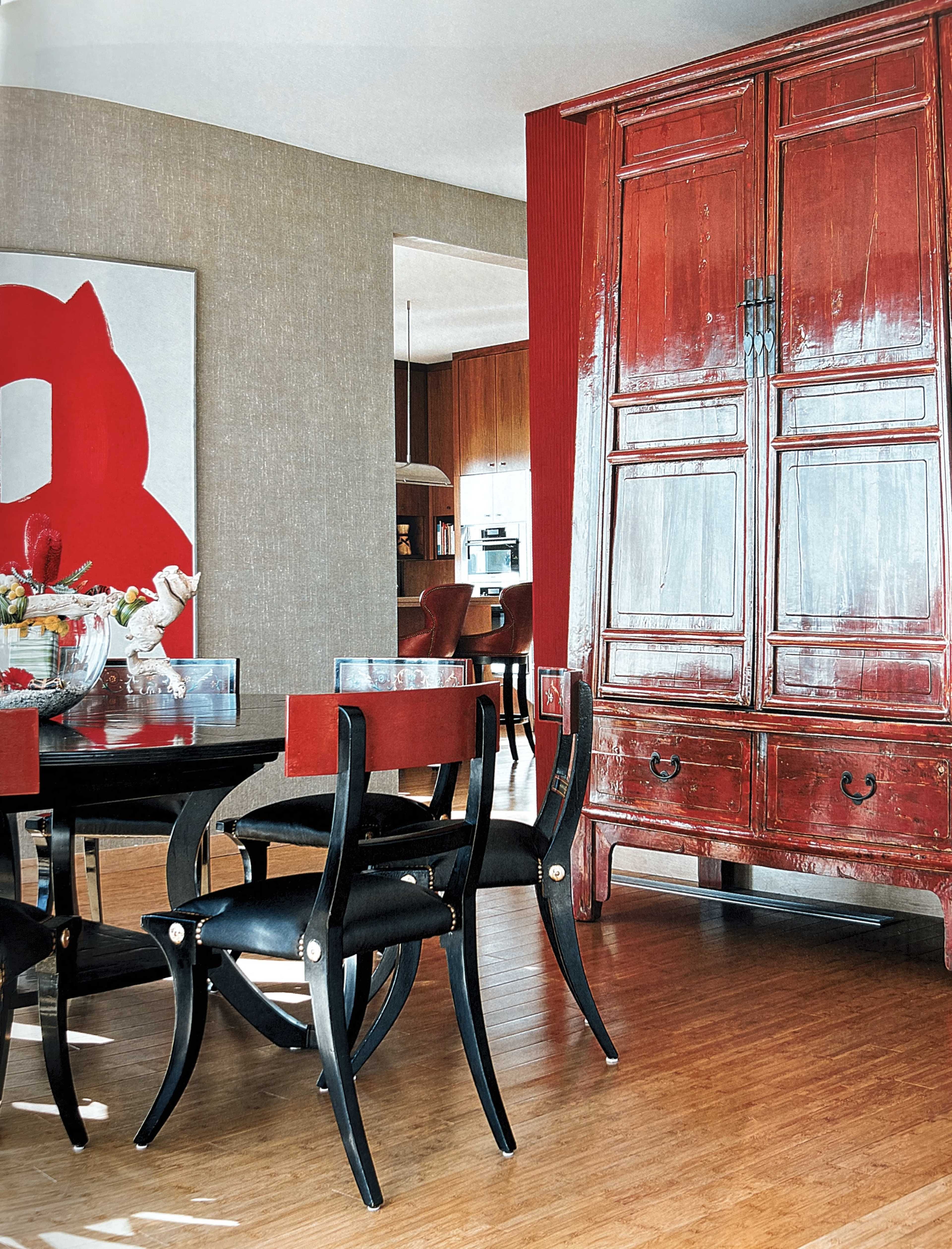 The image shows a modern dining area featuring a round table with black chairs and a large red wooden cabinet against a beige wall, complemented by a piece of abstract artwork.